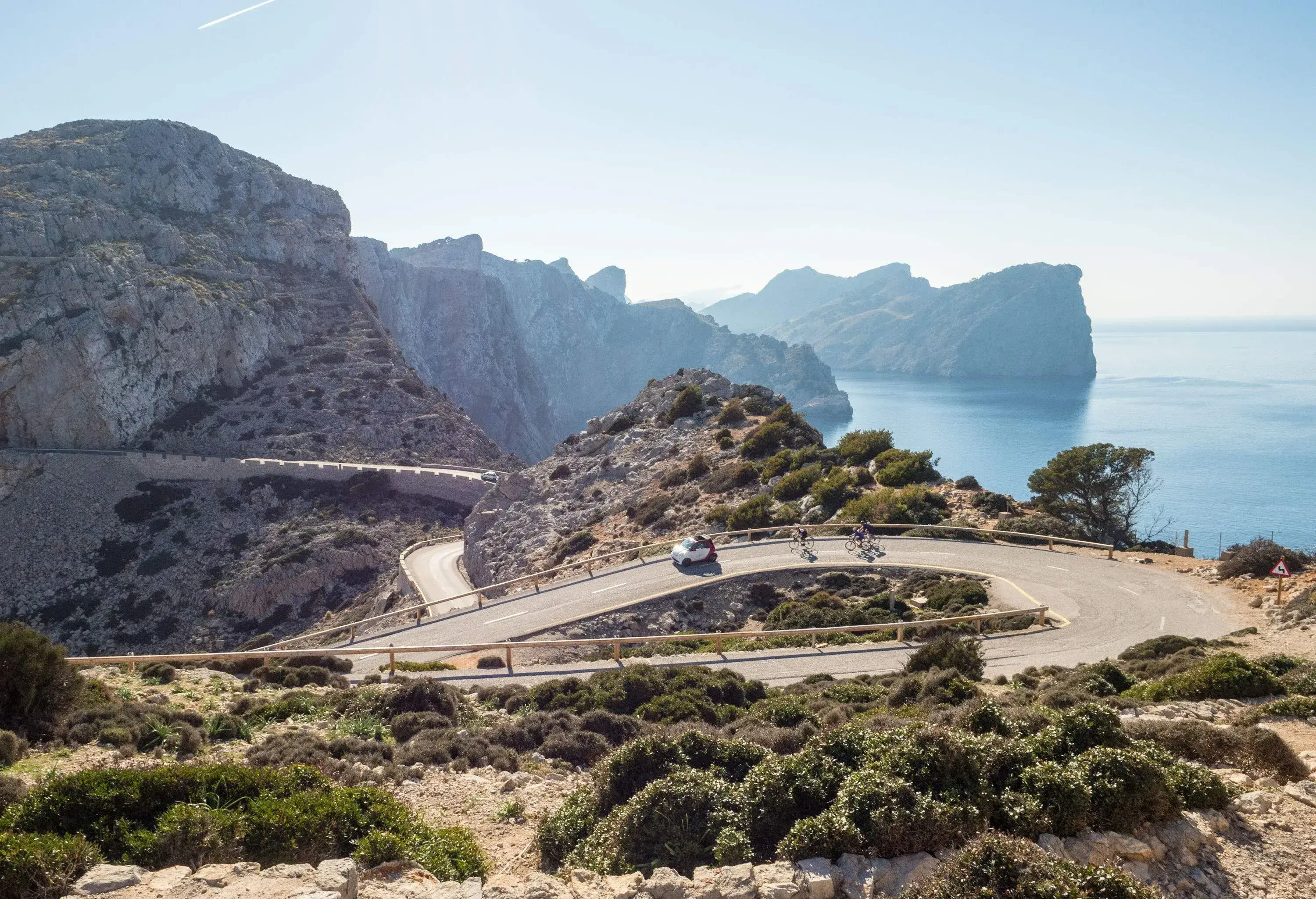 Two cyclists and a car travel down a winding road next to a cliff in a mountainous terrain.
