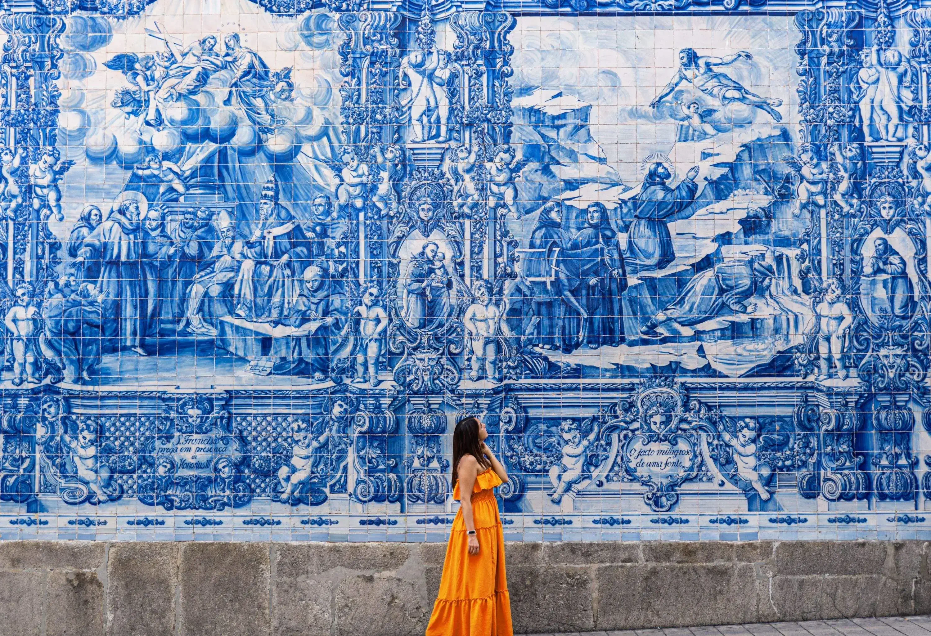 Woman wearing orange dressing looking up in wonder standing against a wall of azulejo tiles