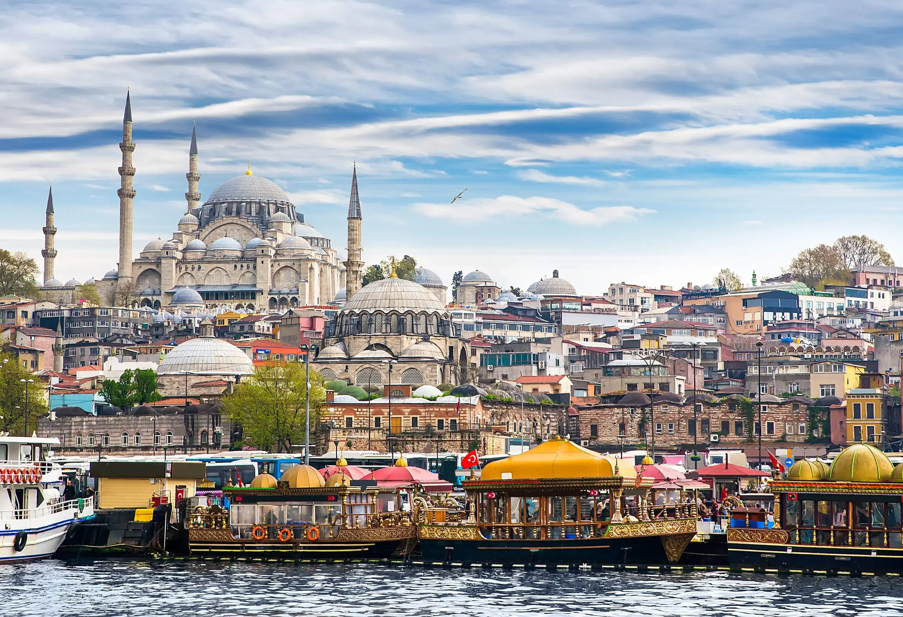 A harbour with anchored ferries with colourful dome roofs against the backdrop of a massive mosque overlooking a cluster of city buildings.