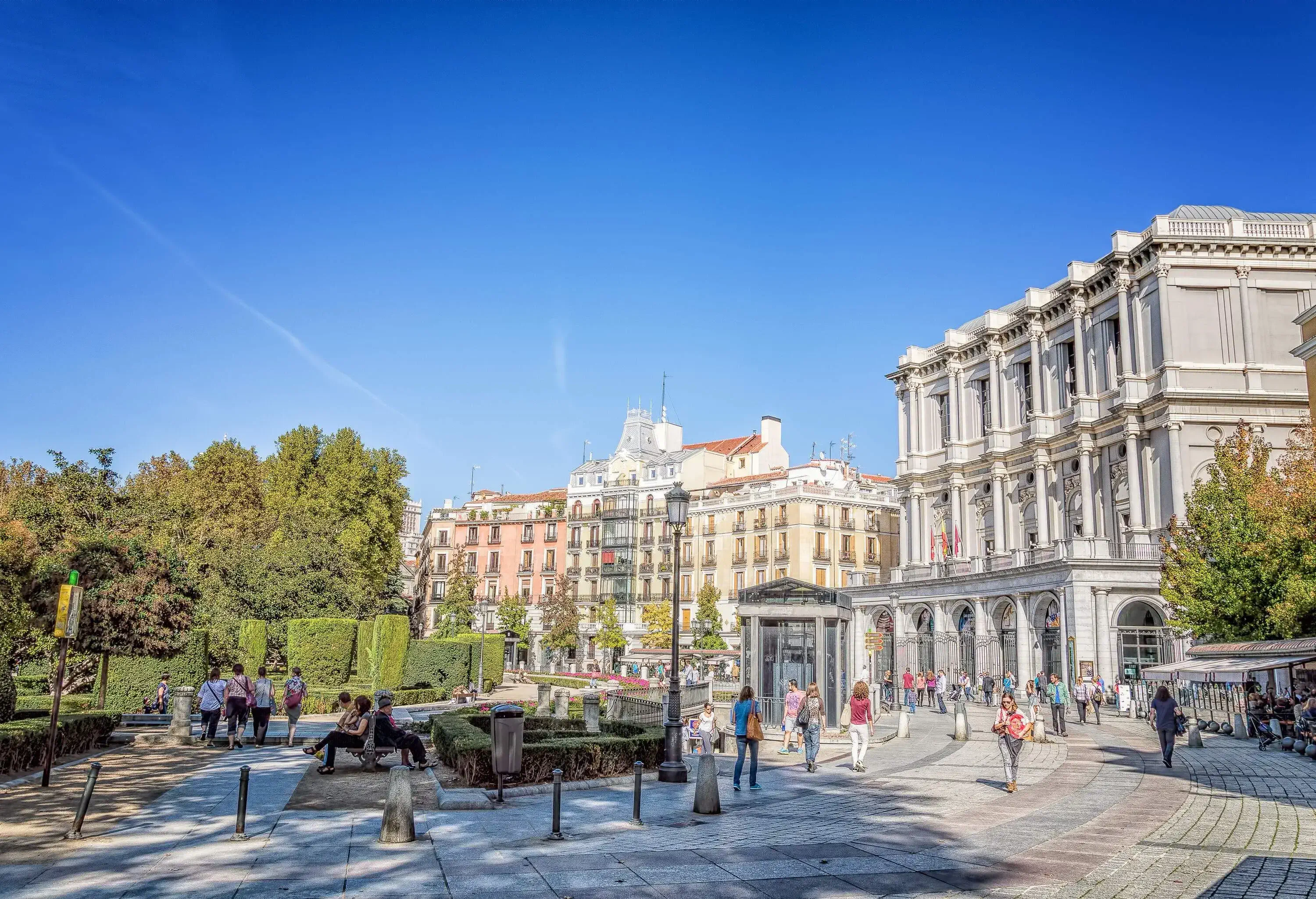 People along the plaza with trees and shrubs with two buildings behind.