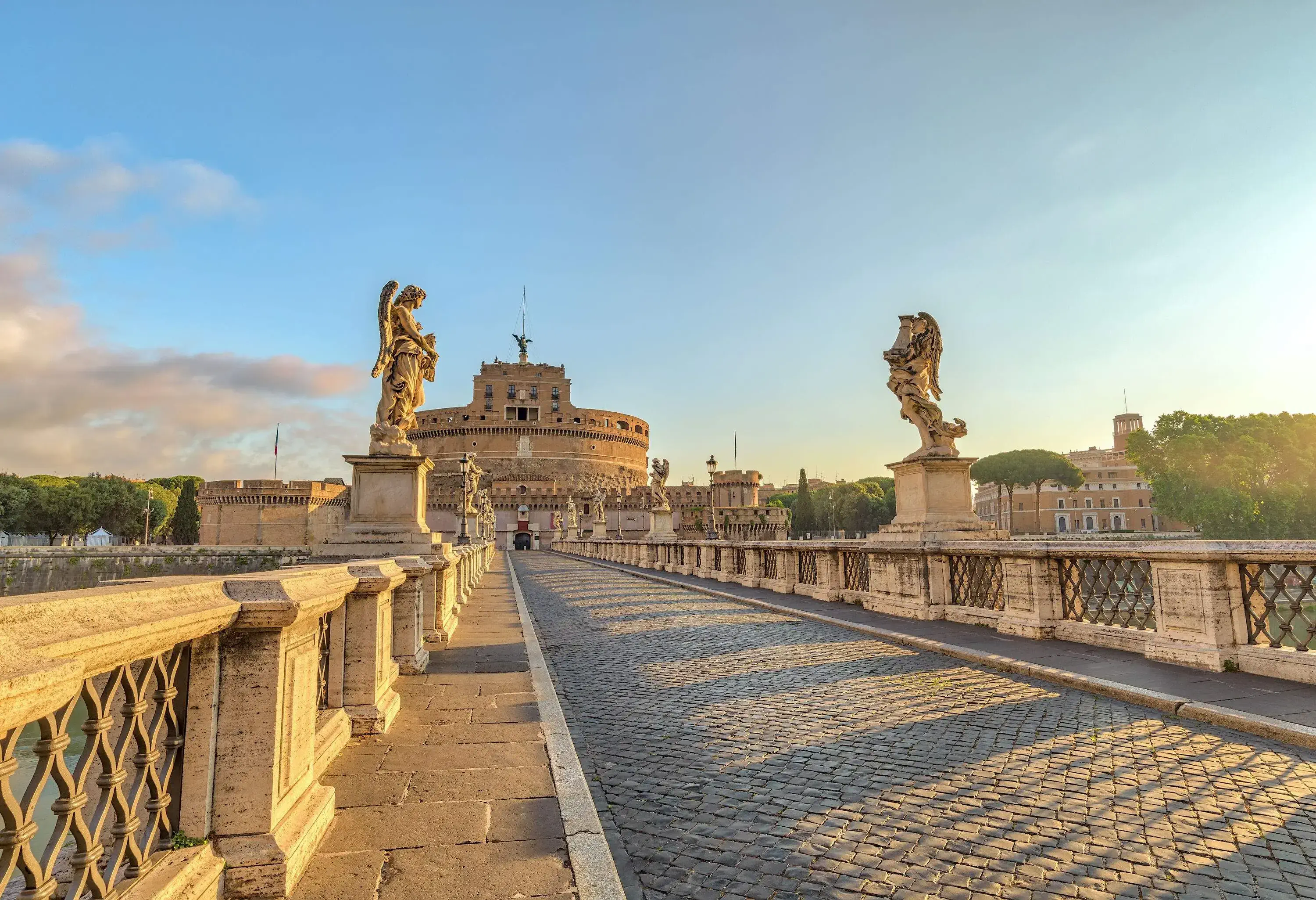 An empty street lined with railings and angel statues towards Castel Sant'Angelo, a cylindrical castle with high walls.
