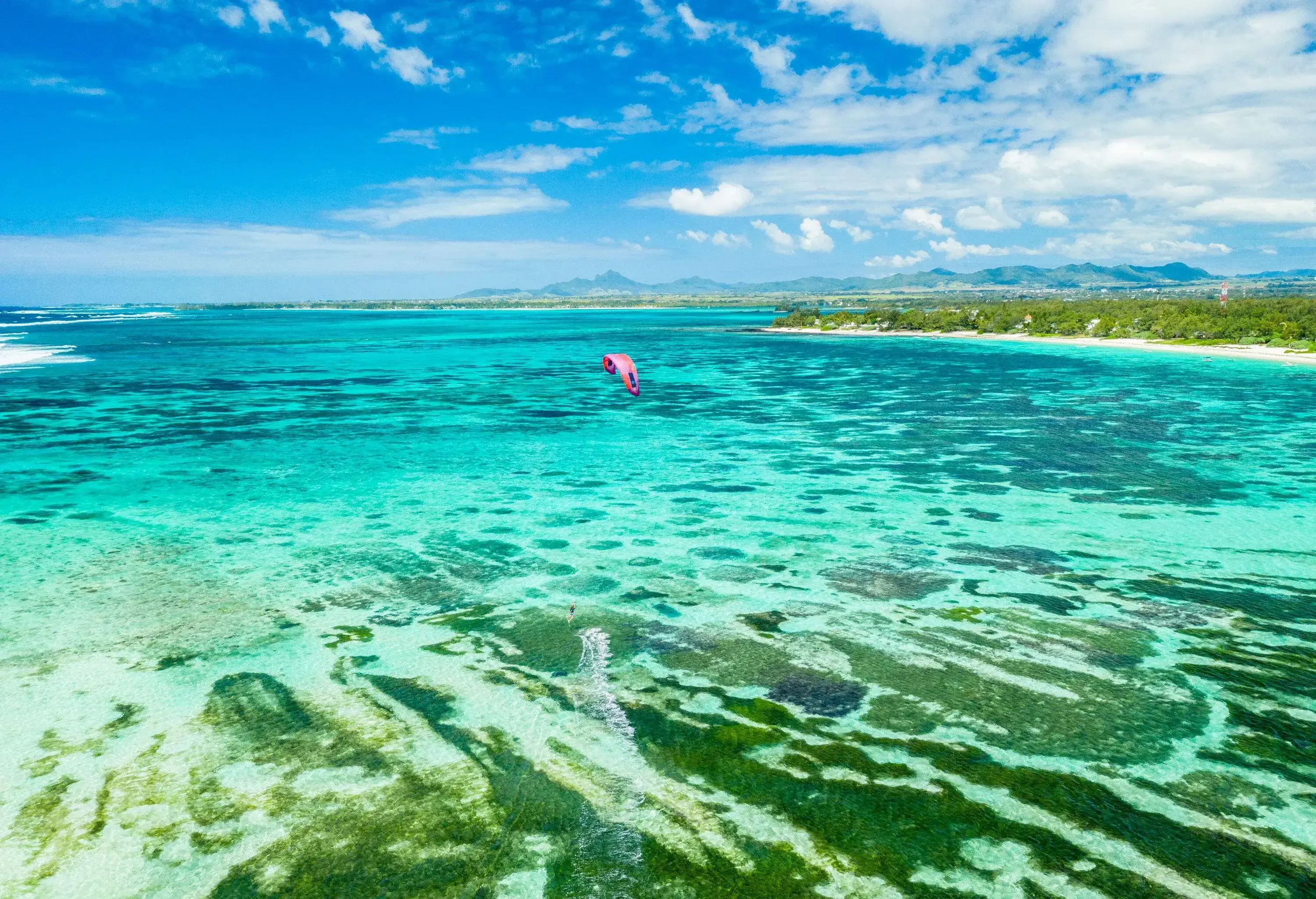 A kite surfer skillfully navigates along a vibrant coral reef, leaving a frothy trail in the crystal-clear turquoise water.