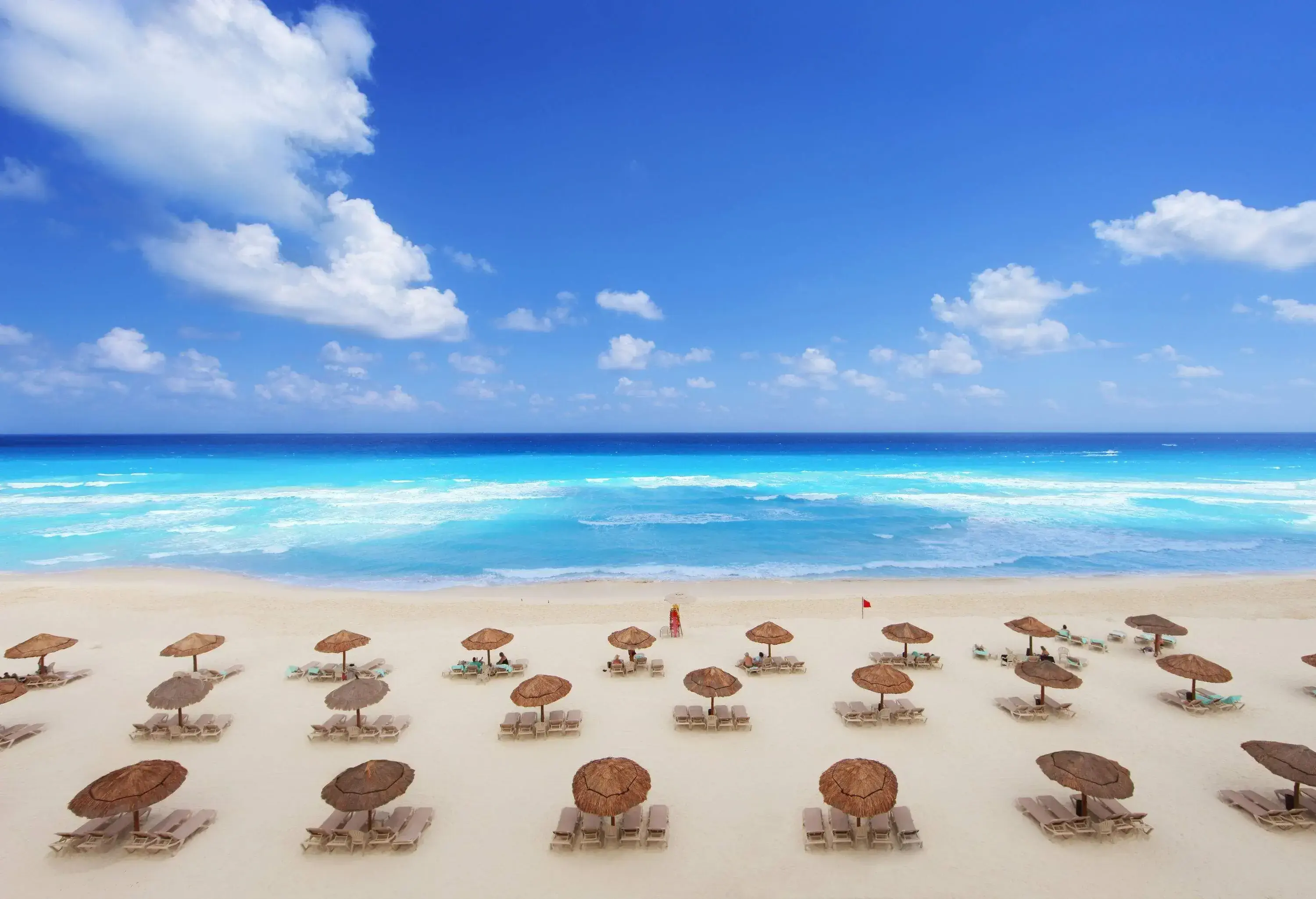 A beach with rows of sunbeds under thatched umbrellas in front of azure waves.