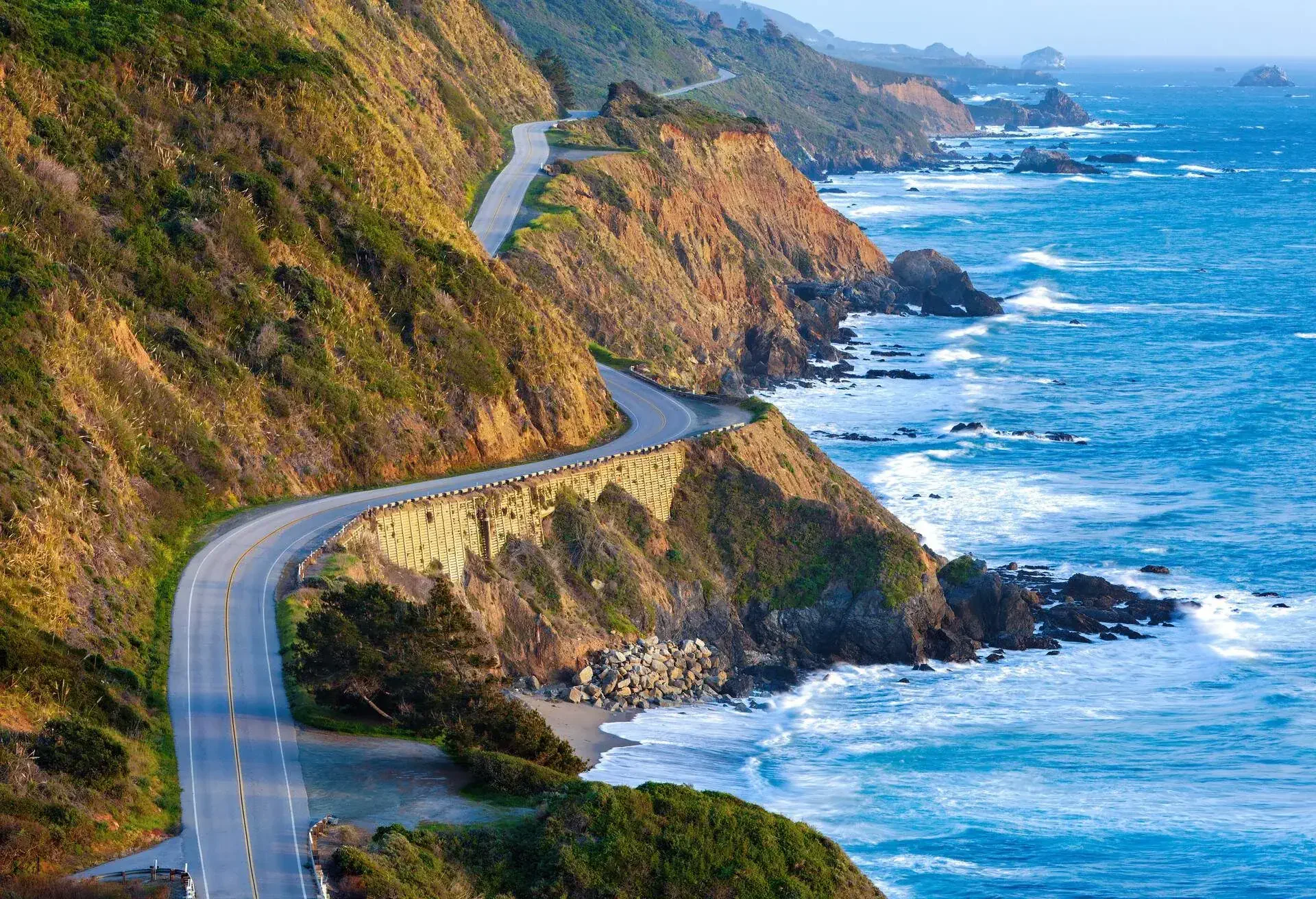 A winding road with views of a rocky beach carved into the faces of high coastal cliffs.