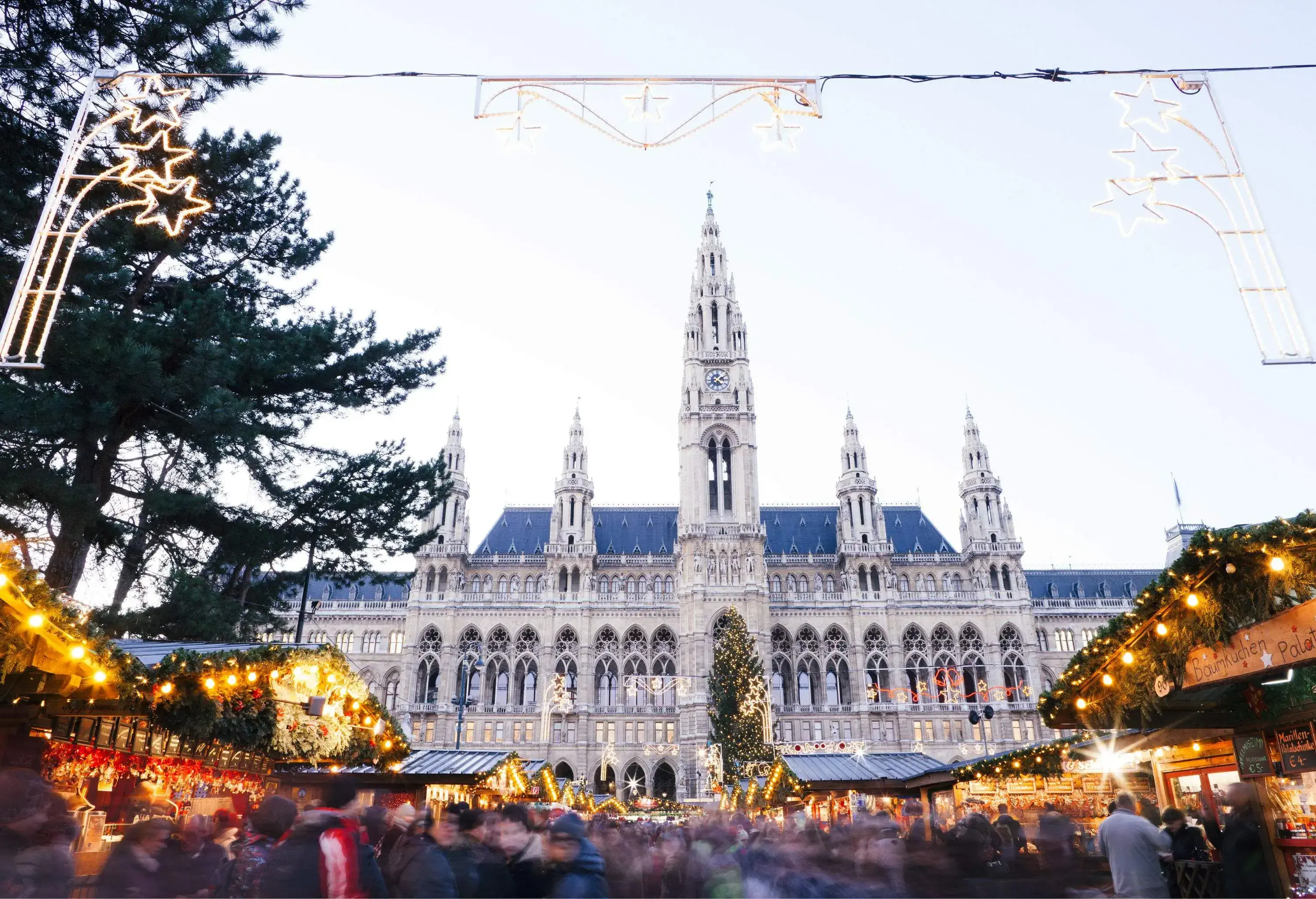 A grand monumental building serving as a backdrop to a Christmas market lavishly decorated with garlands and lights.