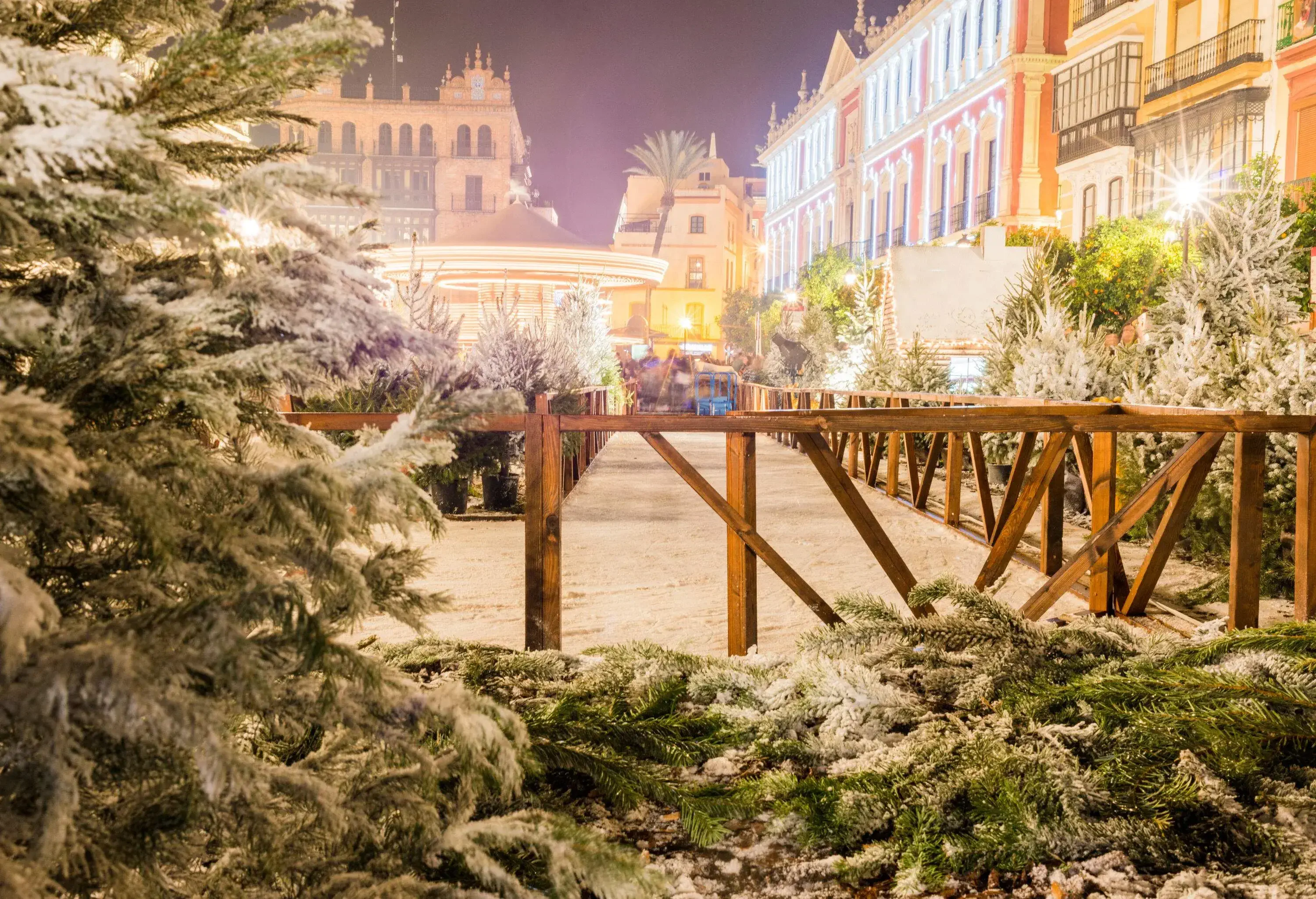 A gated snow path surrounded by frosted pine trees and adjacent buildings.