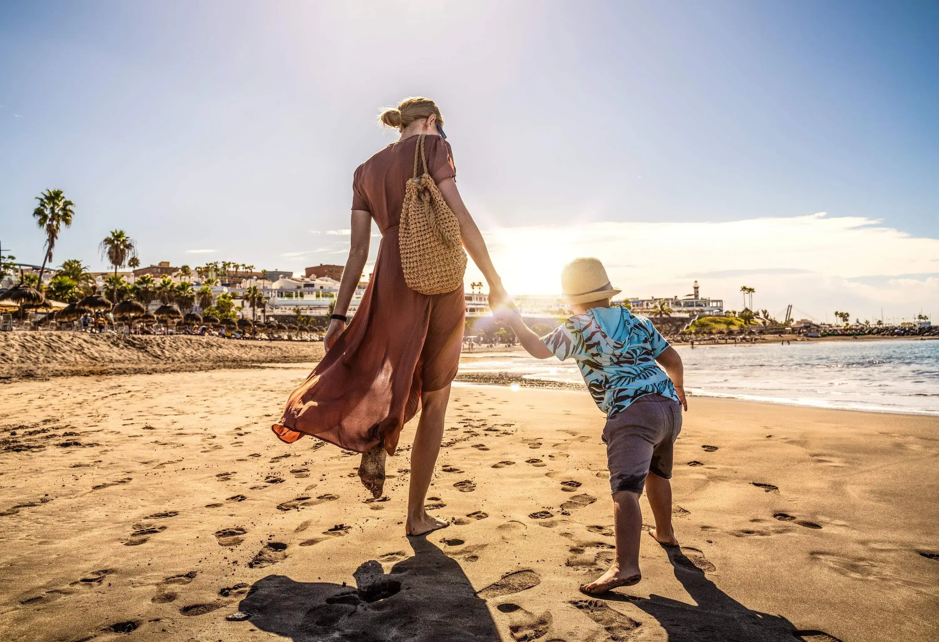 A mother holds his son's hand while walking barefooted on wet fine brown sand under the sun.