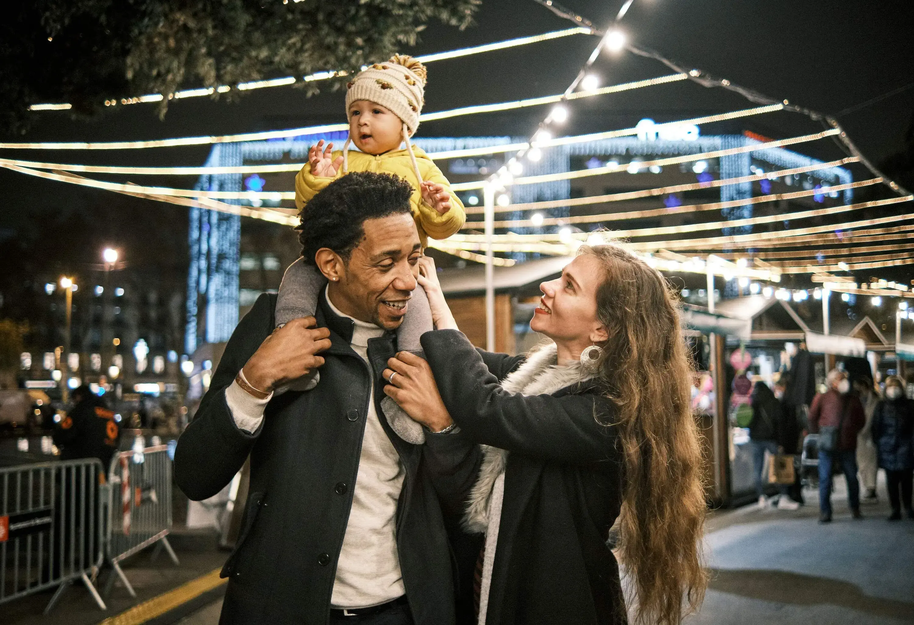 A father with a child on his shoulders and a smiling mother looking at her baby in front of a brightly lit Christmas market.