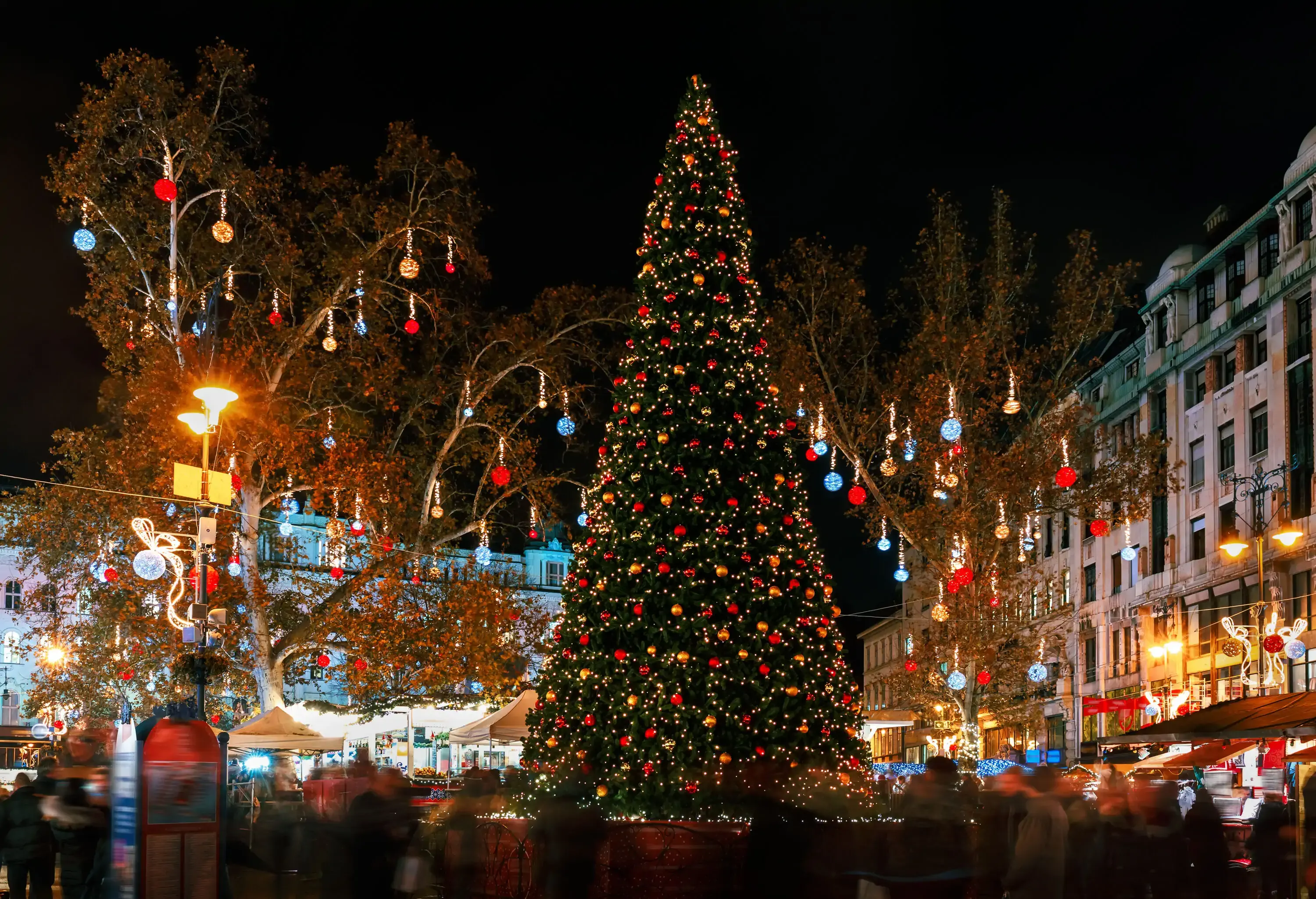 A bustling, brightly lit Christmas market with a tall and colourfully ornamented Christmas tree in the centre.