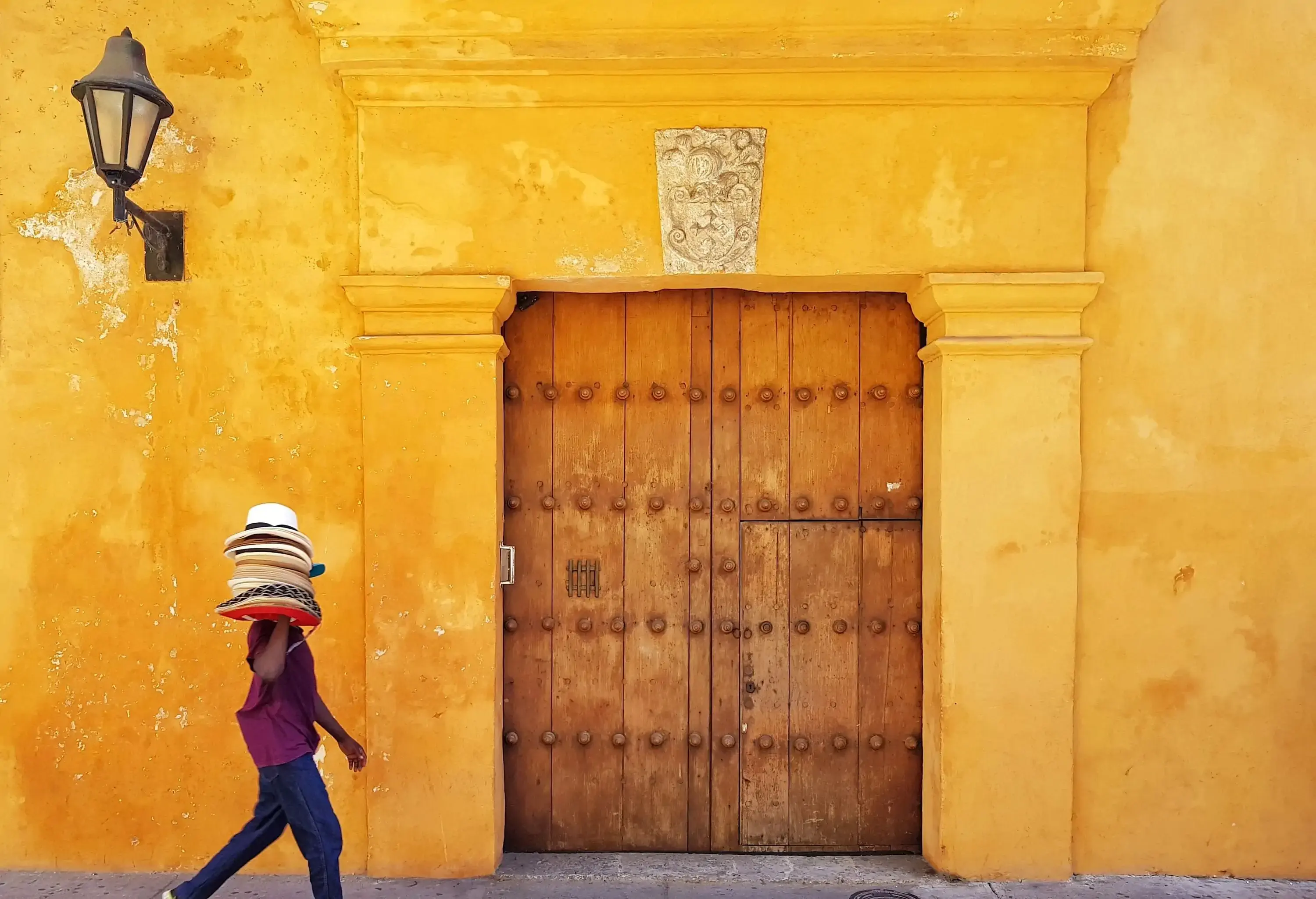 A hat seller gracefully carried merchandise in one hand, passing by a charming yellow building with a large wooden door.