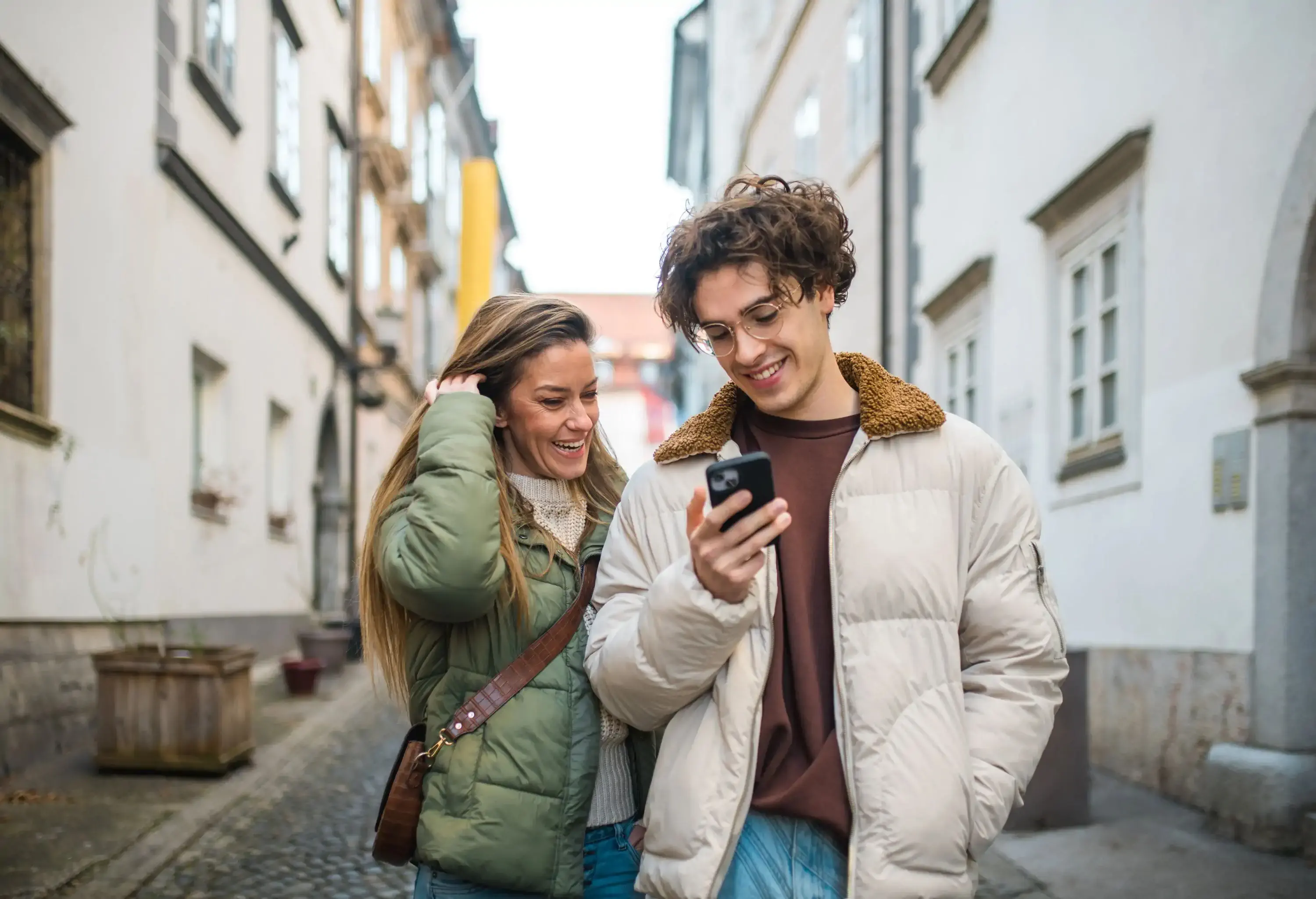 Happy Caucasian woman with her young adult son standing in an urban setting engaging in a video call. Both wear casual autumn jackets, sharing a joyful moment with their mobile device.
