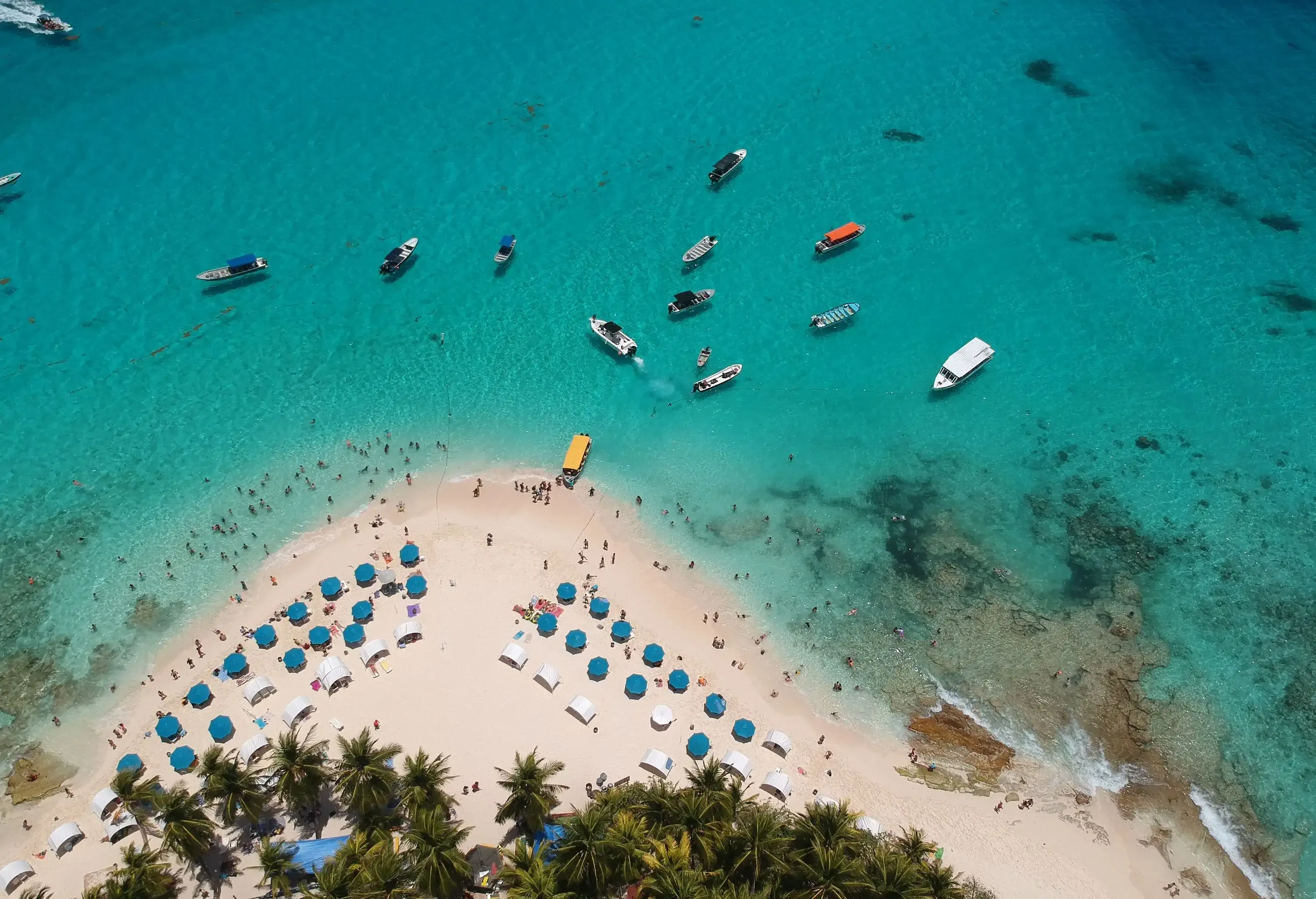 A not-so-crowded tranquil beach with rows of blue and white umbrellas and several boats moored on the crystal clear turquoise water. 
