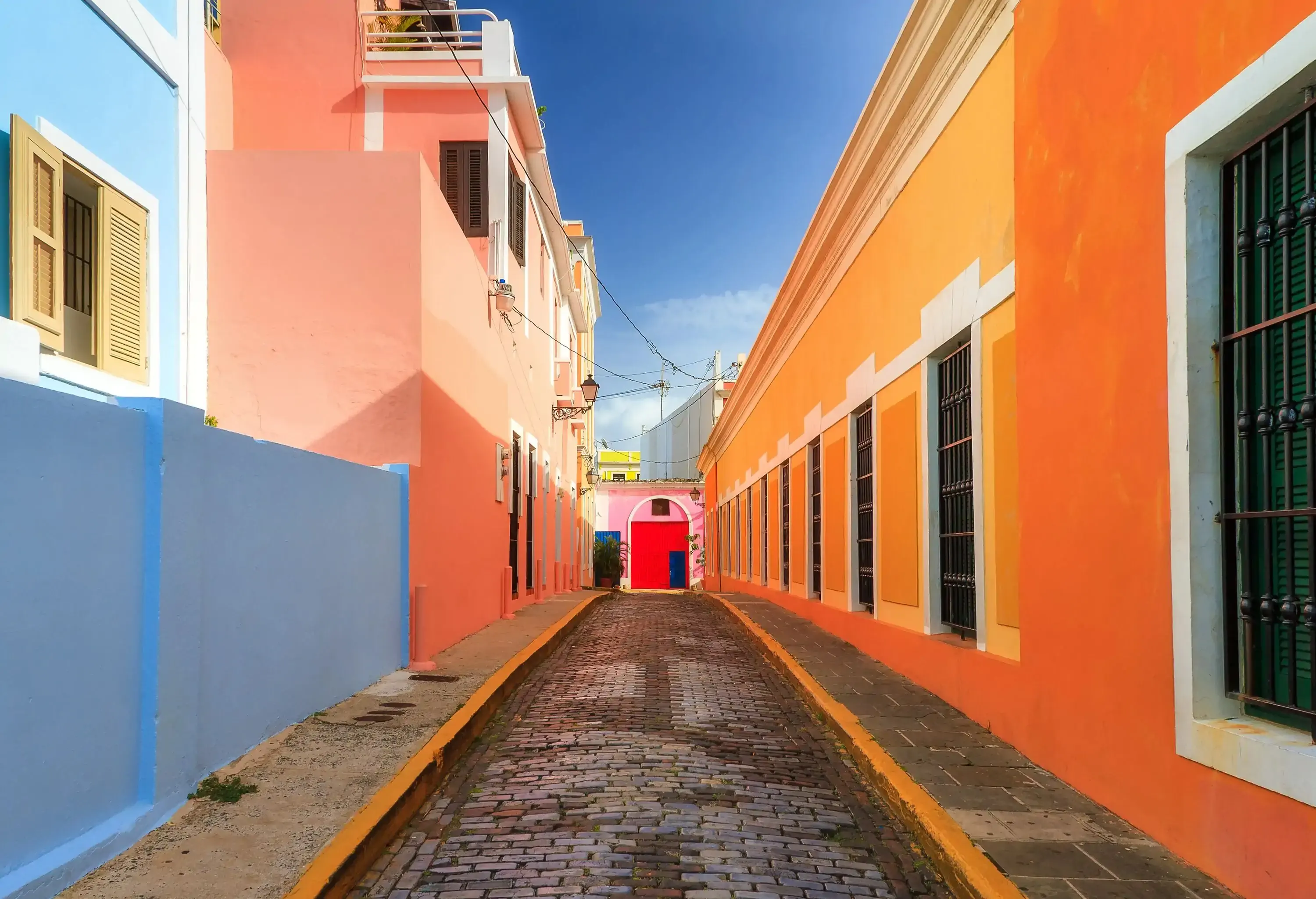 Buildings of various colours lining both sides of a paved alleyway, their rooftops framing the blue sky.