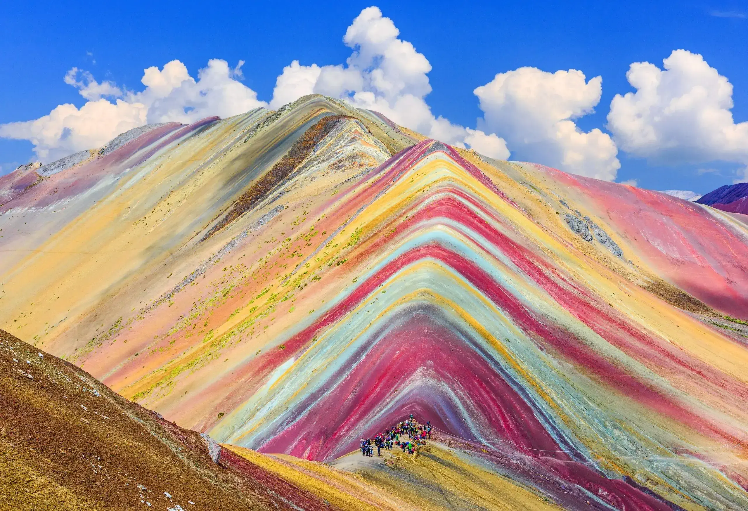A group of hikers stand on a ridge as they enjoy the views of a colourful mountainous terrain.