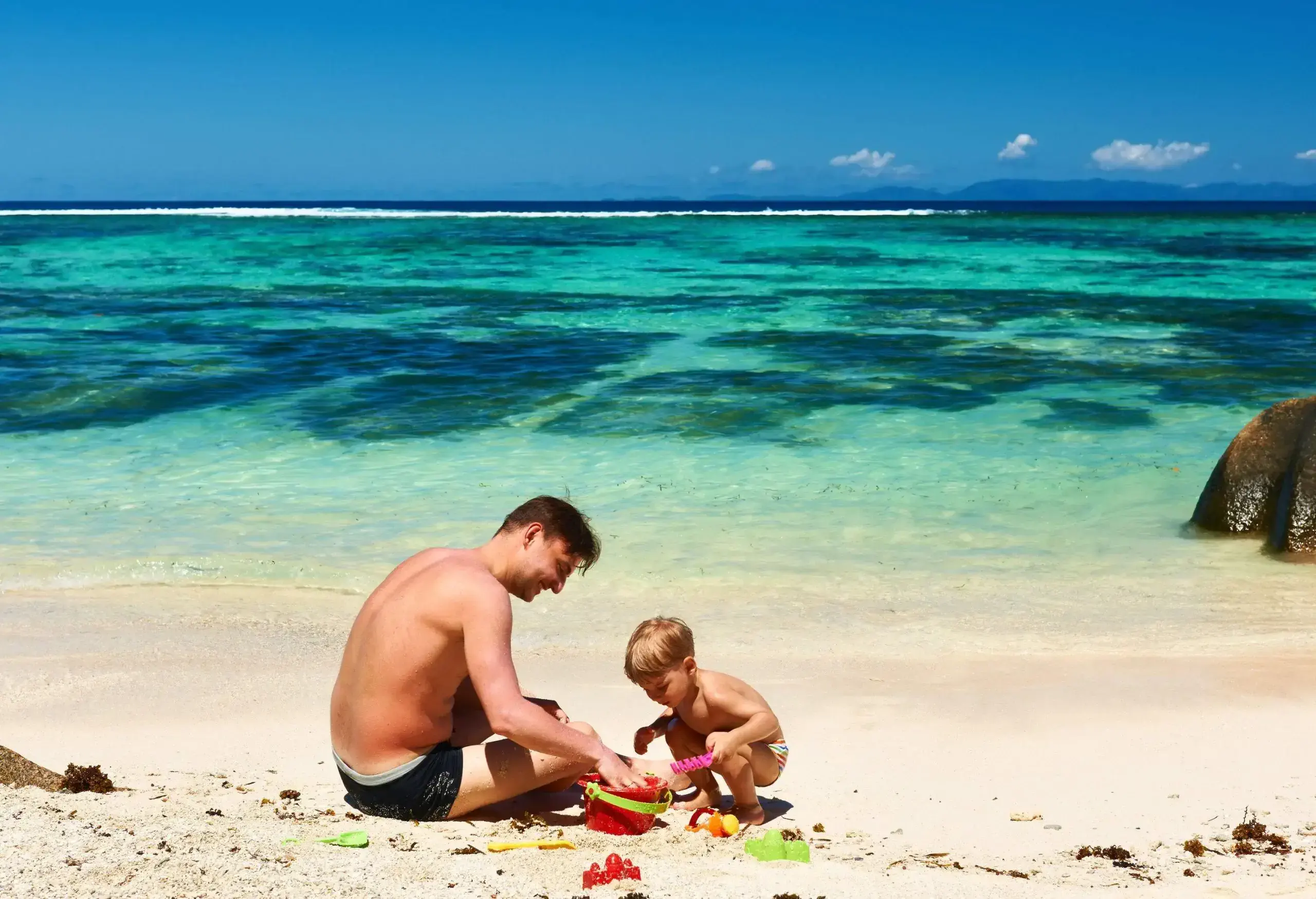 A man and a kid, both in swimming trunks, enjoy playing with sand on a beach with turquoise waters.