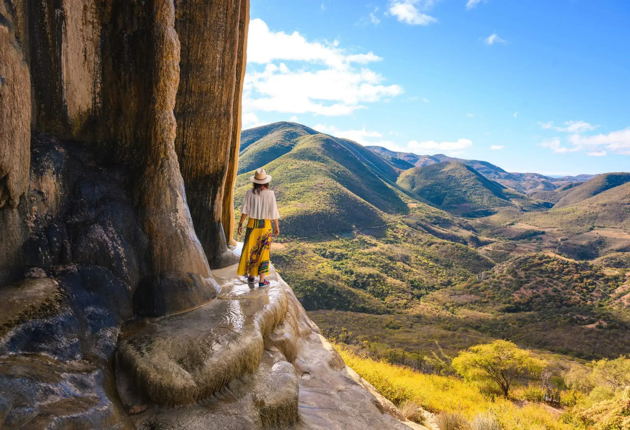 A woman stands on a steep natural rock overlooking lush green hills.