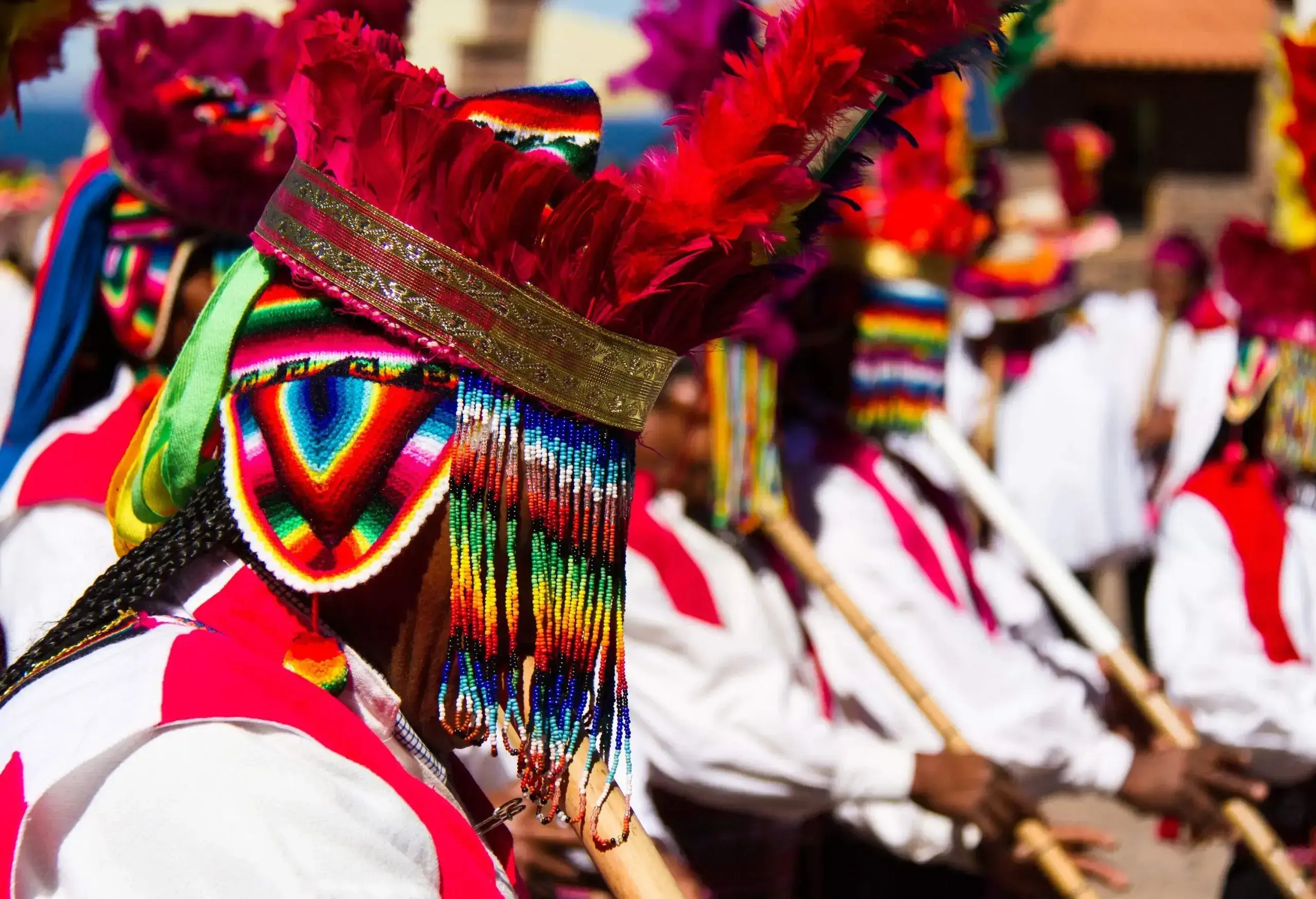 A man in traditional attire wears a headdress with red feathers and colourful hanging beads while playing a wooden flute.