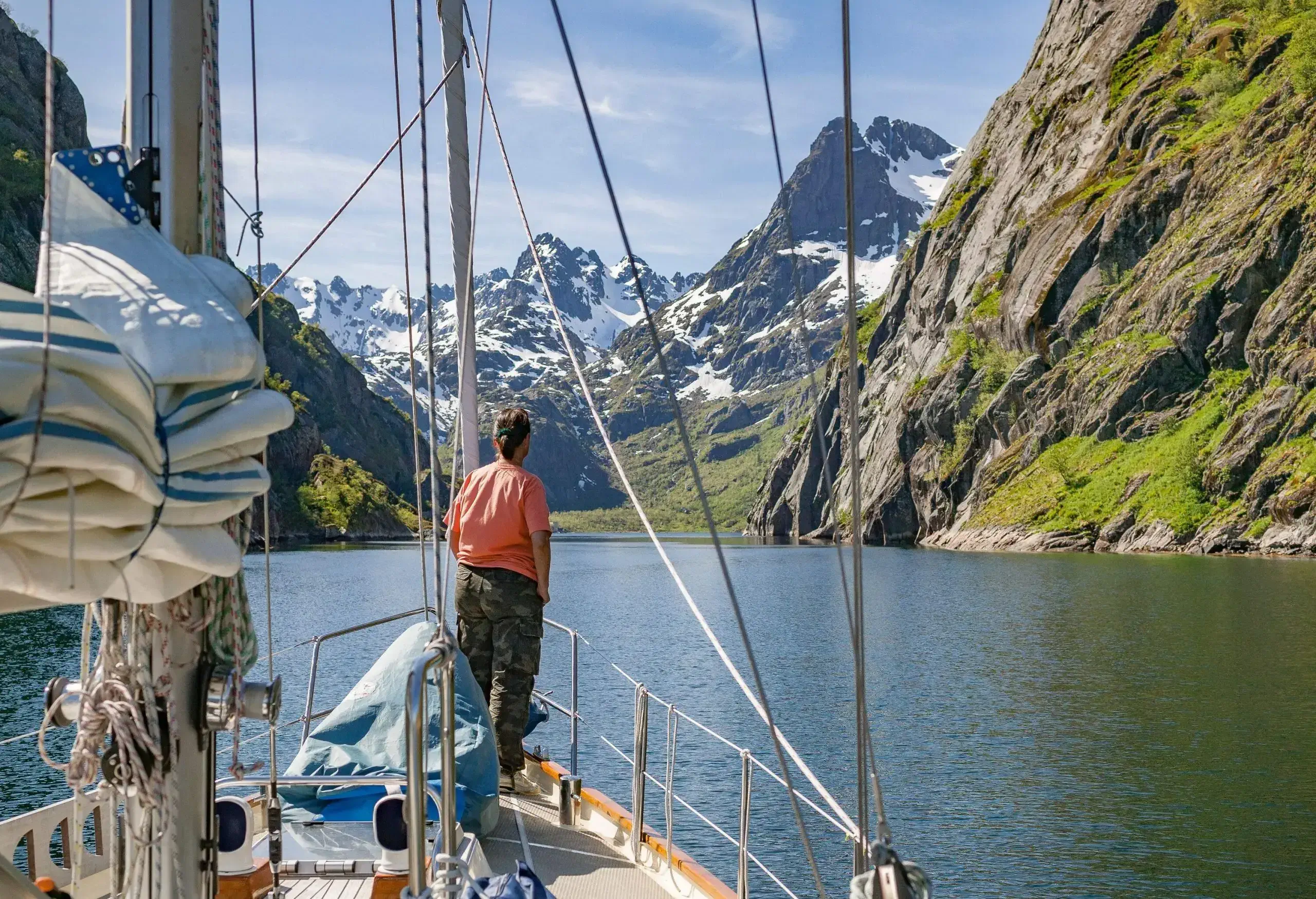 A person stands on a sailboat's front deck gazing at the vast lake and snow-capped mountain views.