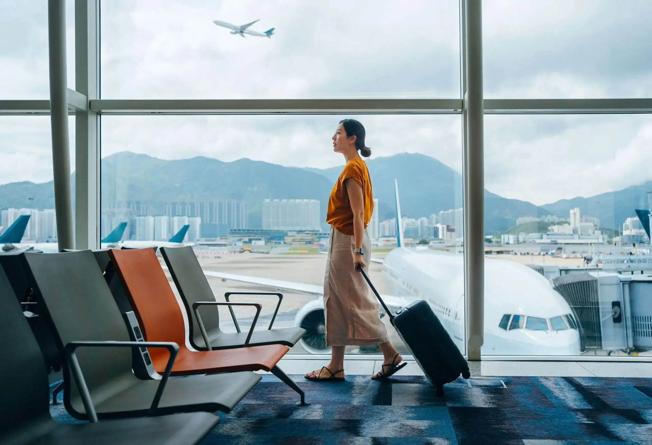 Young Asian woman carrying suitcase, walking by the window at airport terminal. Young Asian female traveller waiting for boarding at airport. Business travel. Travel and vacation concept
