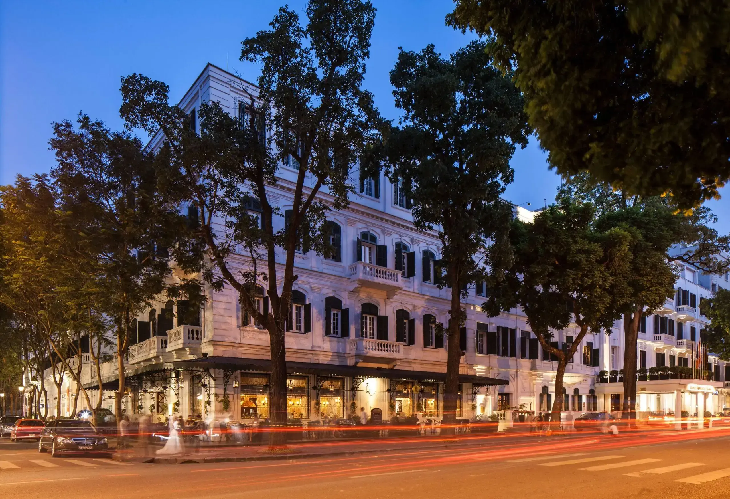 A white building with multiple windows along a busy tree-lined intersection.
