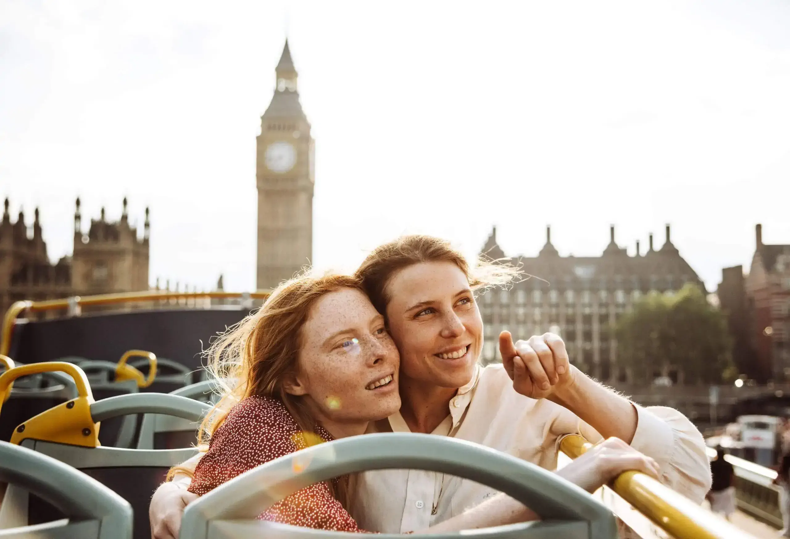 Female lovers sit and hug on an open-top bus while admiring the cityscape.