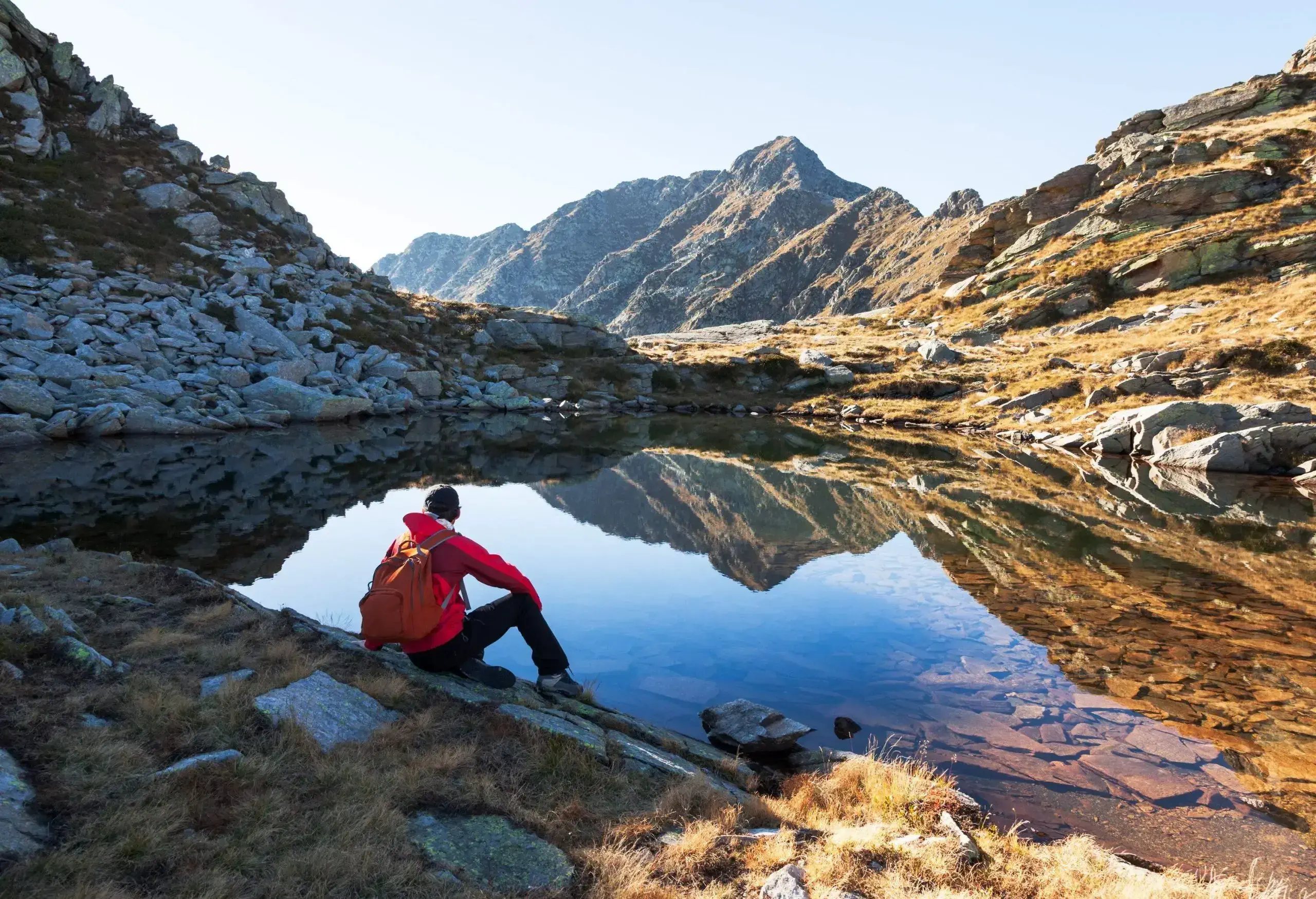 A man sitting on the slope side in front of a clear lake surrounded by rock mountains.