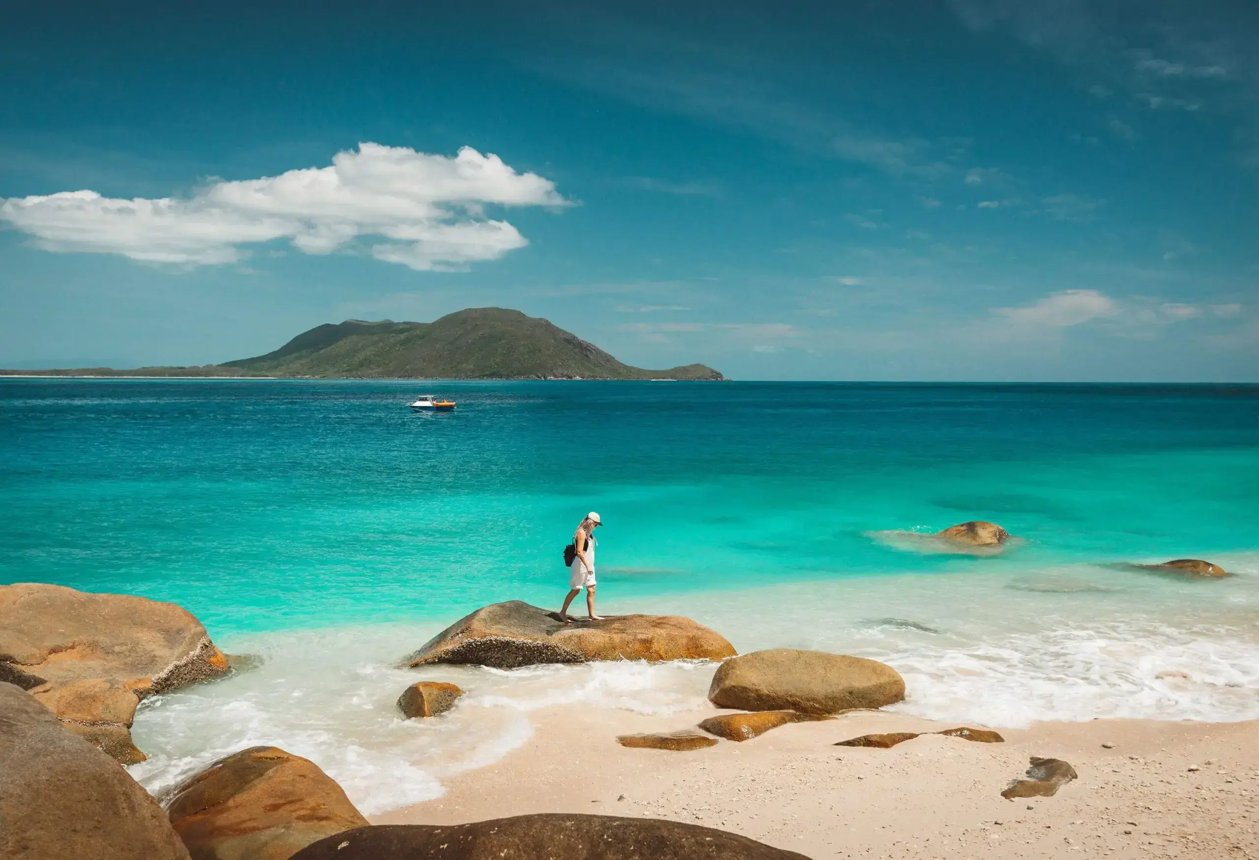 A lone blonde lady stands on a rock on the sandy tranquil turquoise beach with a mountain emerging from the sea in the distance.