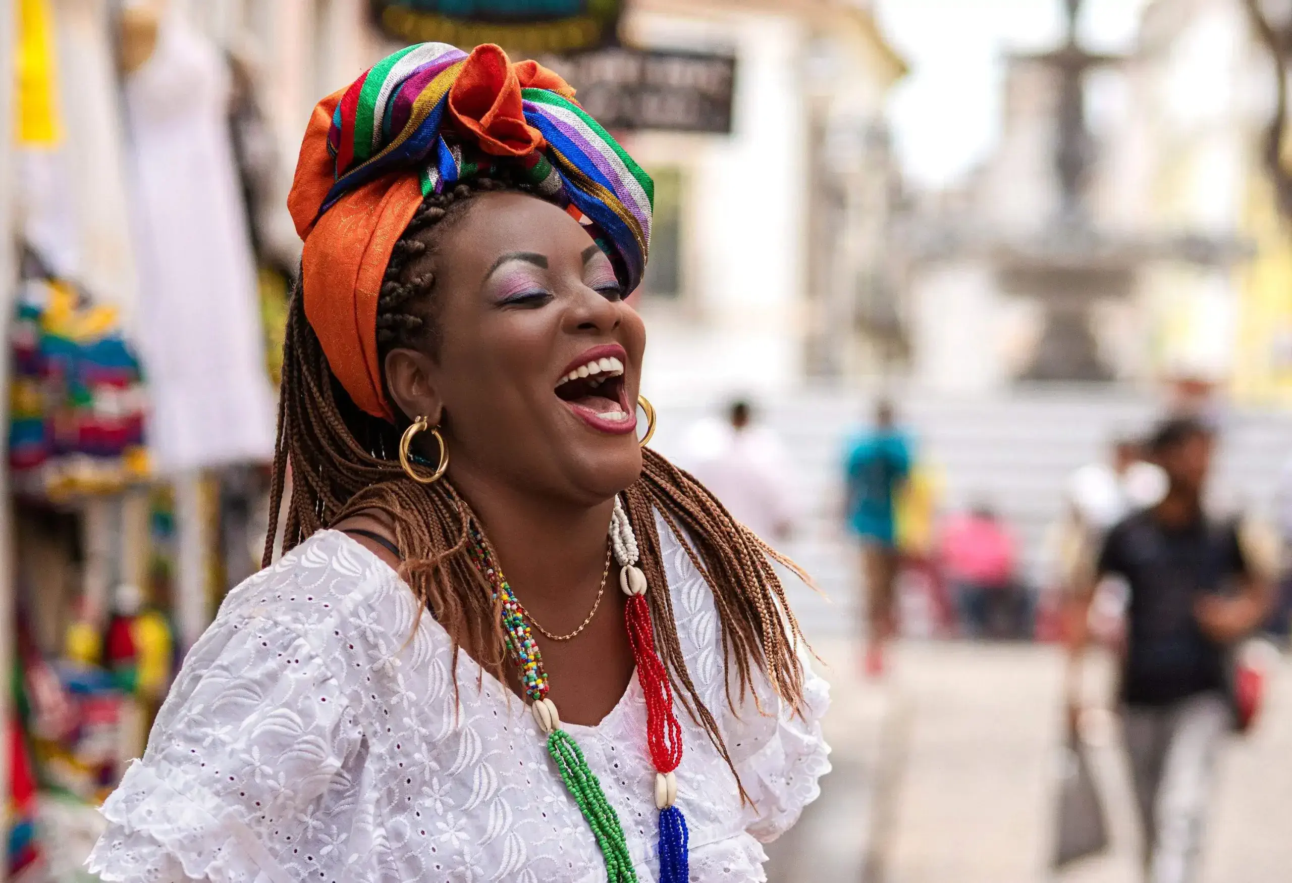 A woman laughing while wearing a white top and a colourful scarf wrapped over her braided hair.