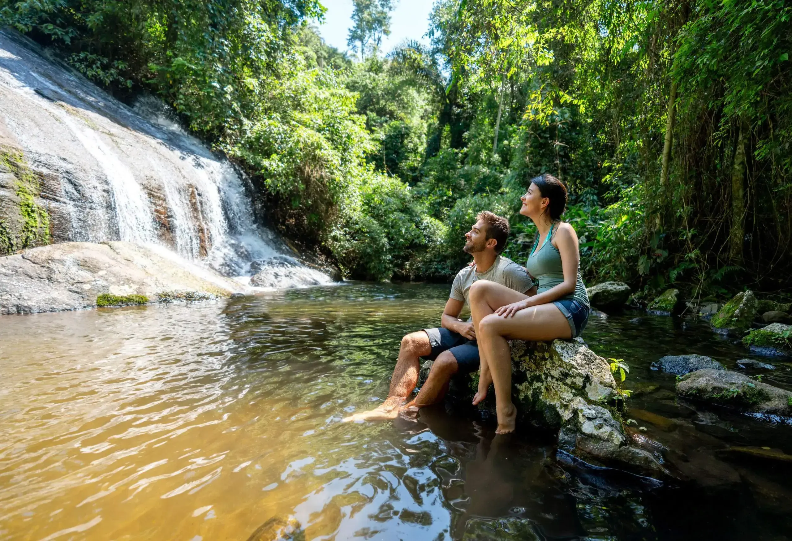 A happy couple sits on a boulder in a river while looking at the waterfalls in the forest.