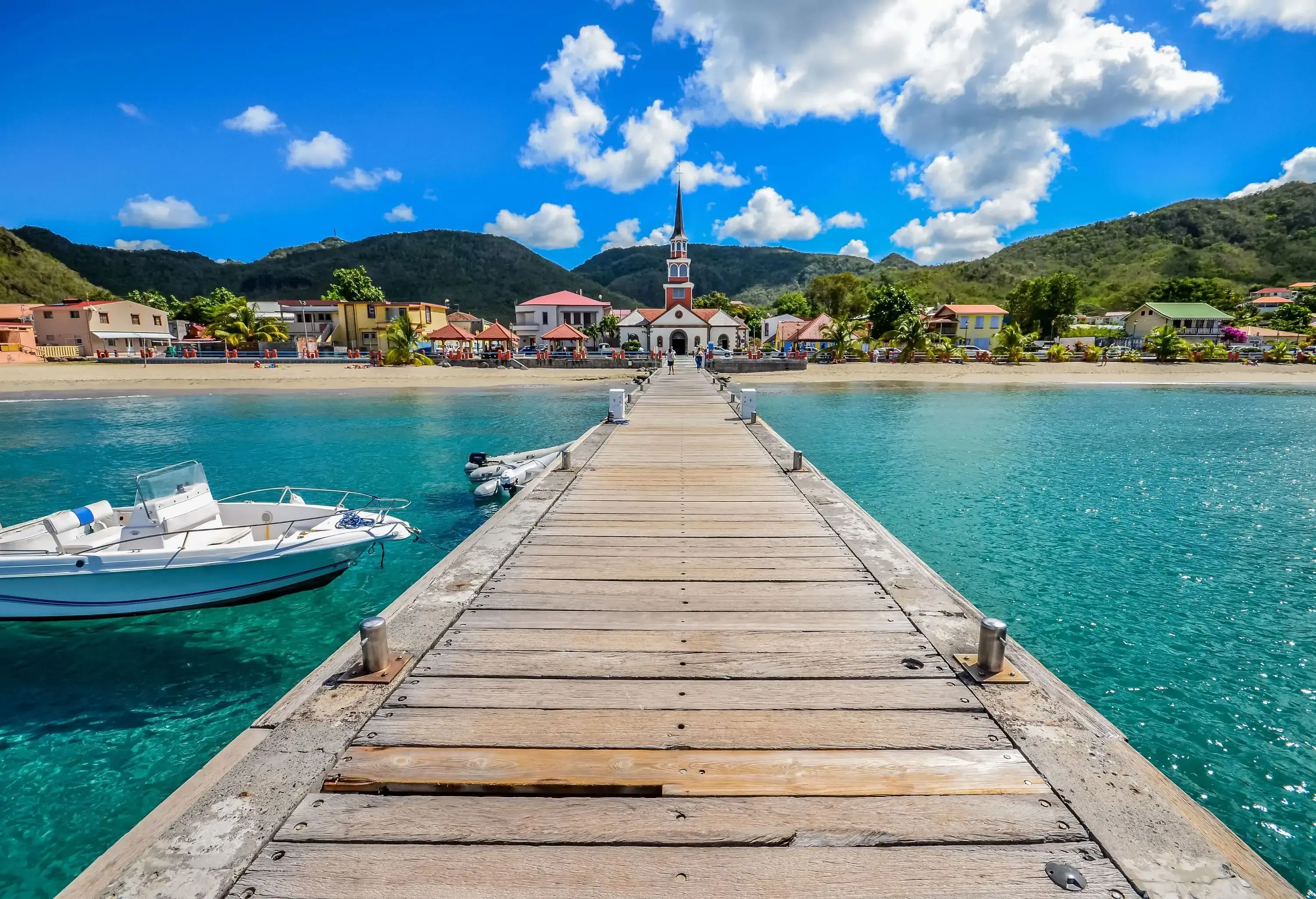 A wooden footpath extends over the turquoise sea, leading towards a shore adorned with beach houses and a church featuring a tall bell tower topped with a spire, all set against a backdrop of forested mountain ranges.
