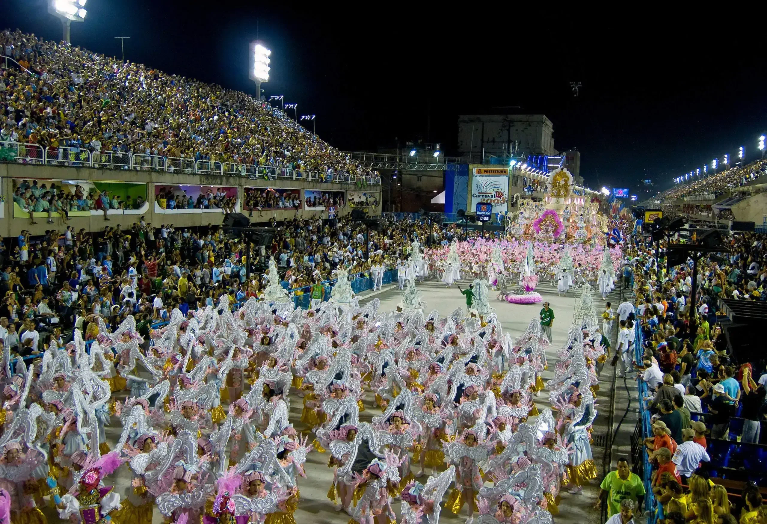 People dressed in stunning white and gold costumes joyously parade through the streets, while crowds of enthusiastic onlookers line the sides, eagerly watching from bleacher-type structures.
