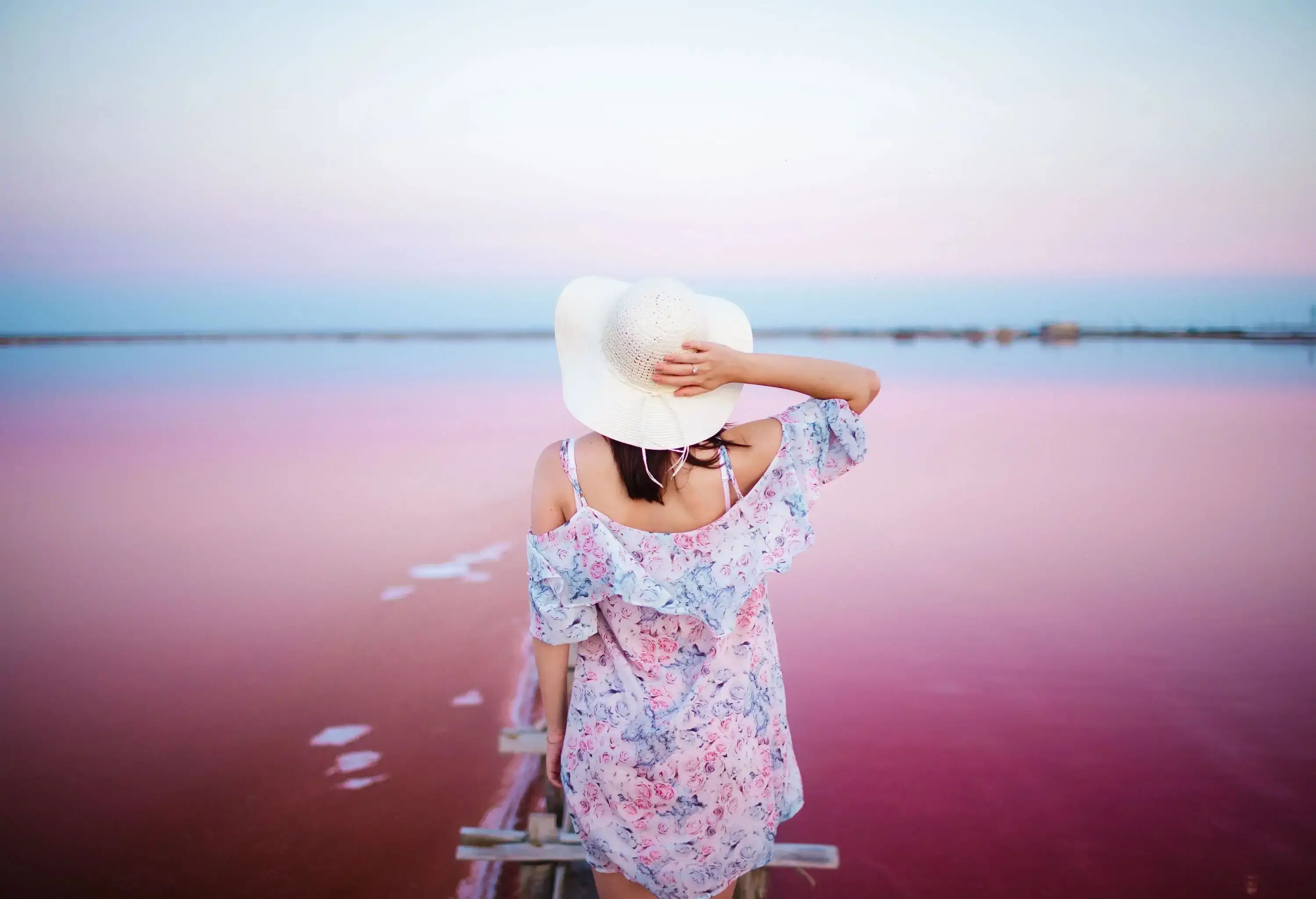 A woman in a floral dress and hat standing ang gazing over at a pink lake.