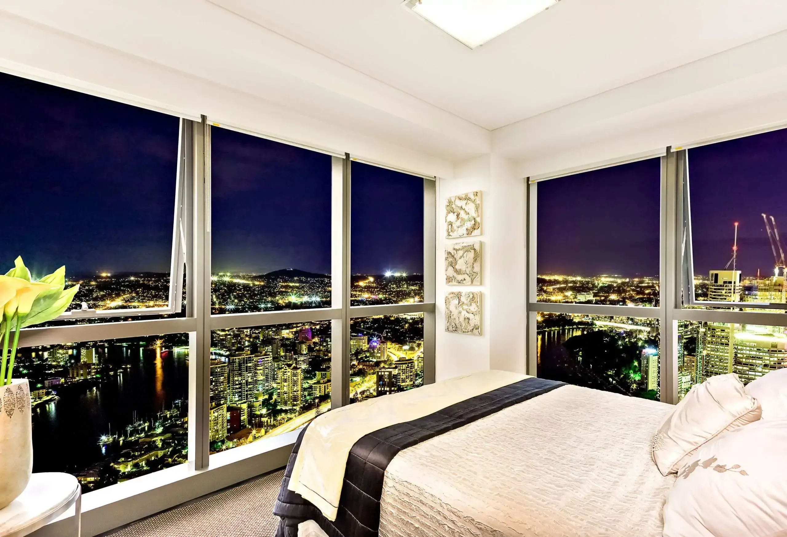 A bed in a room with glass windows that overlooks a dazzling cityscape at night.