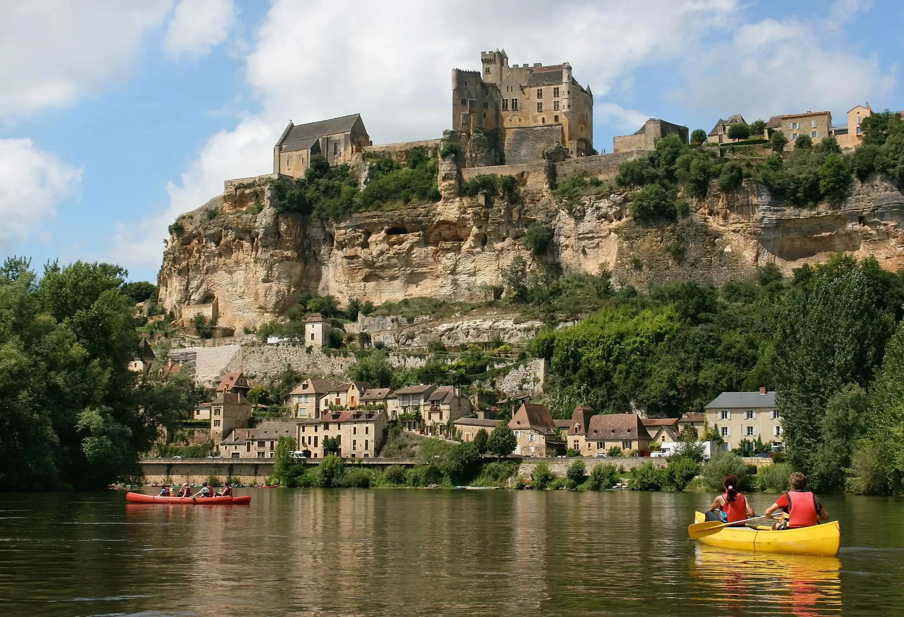 People canoeing across a river with old houses on its bank and an ancient castle on a cliff.