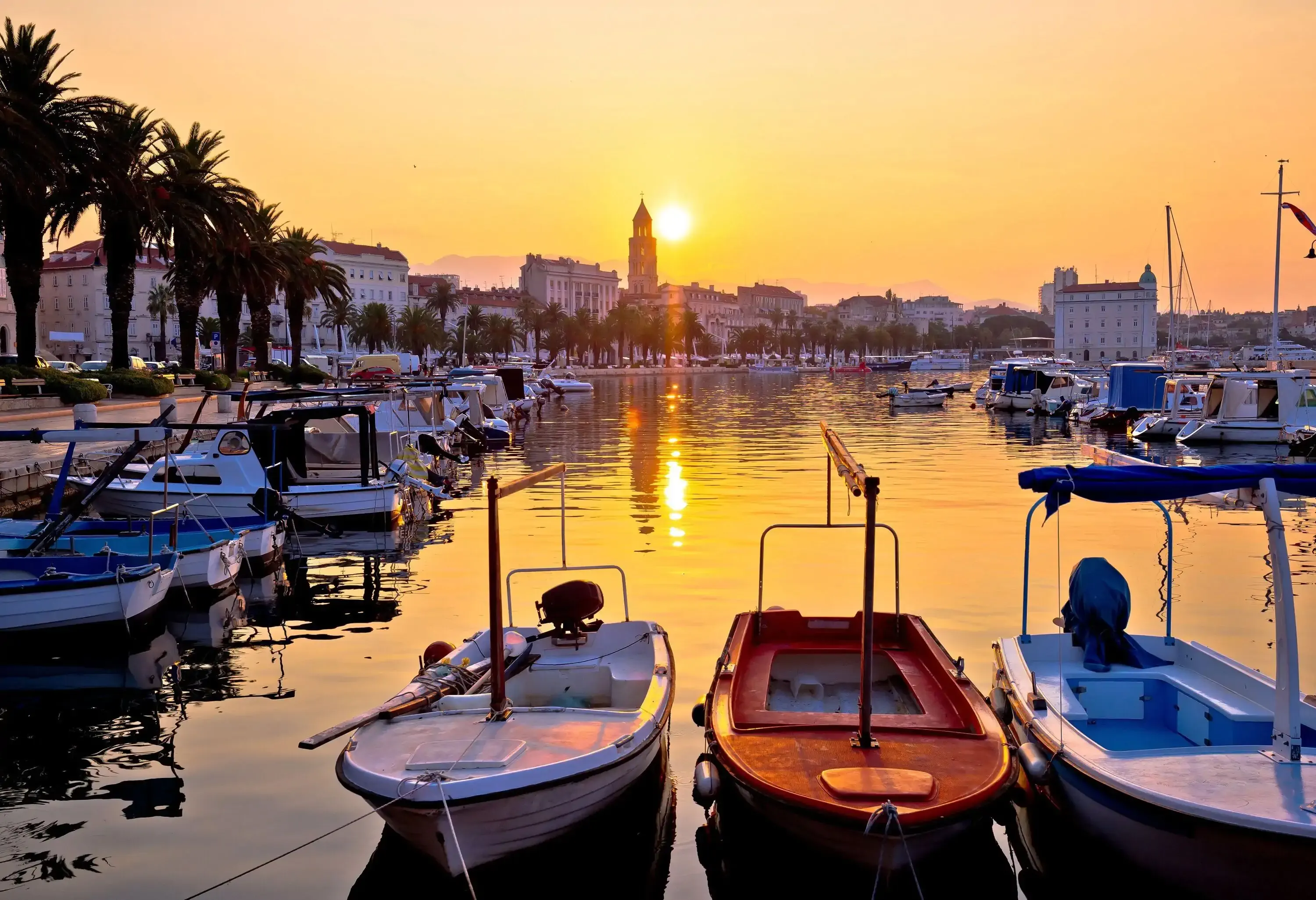 The sun rises above a tower lined with tall buildings shining through the harbour with numerous boats.