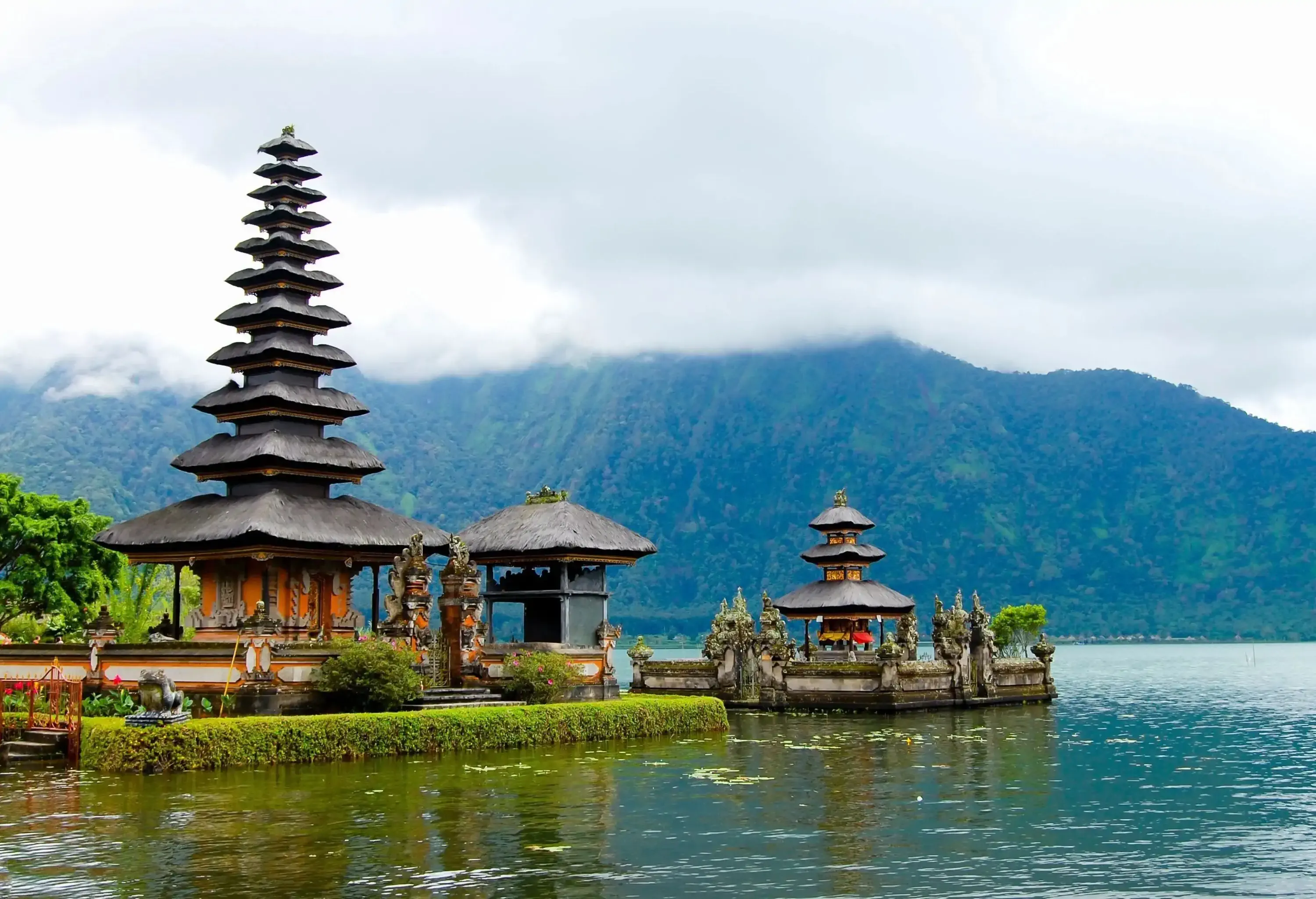 A pagoda-like with the thatched roofs temple complex on the shore of a lake with a mountain range and cloudy sky in the background.