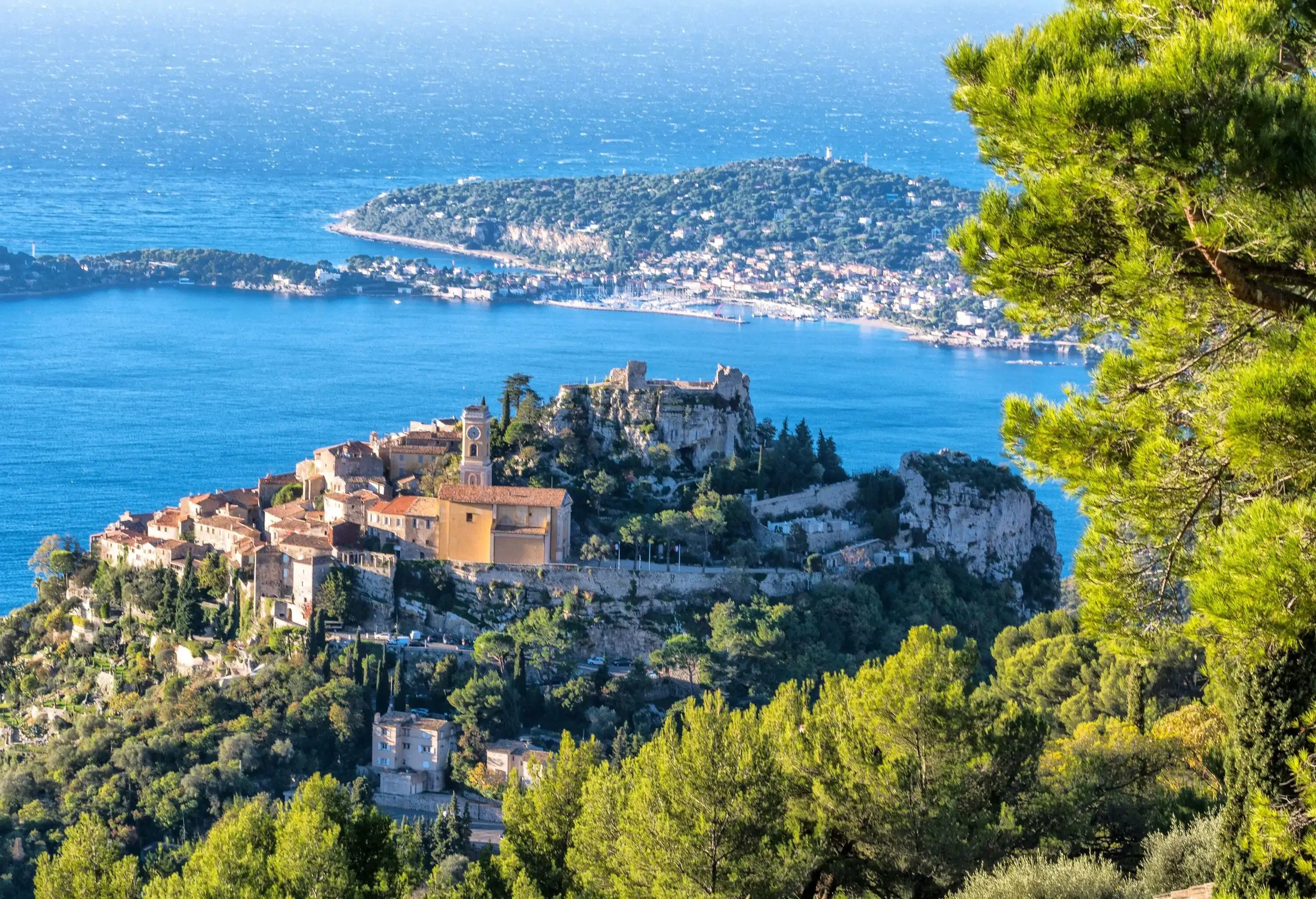 A medieval village on a hilltop surrounded by trees and overlooking the blue sea.