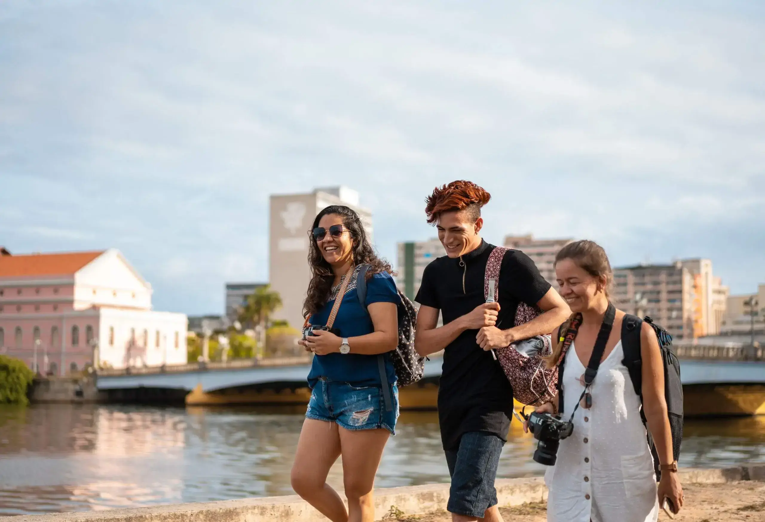 A group of cheerful tourists, donning backpacks, stroll along the scenic Capibaribe River, their smiles shining bright as the picturesque background fades into a gentle blur.