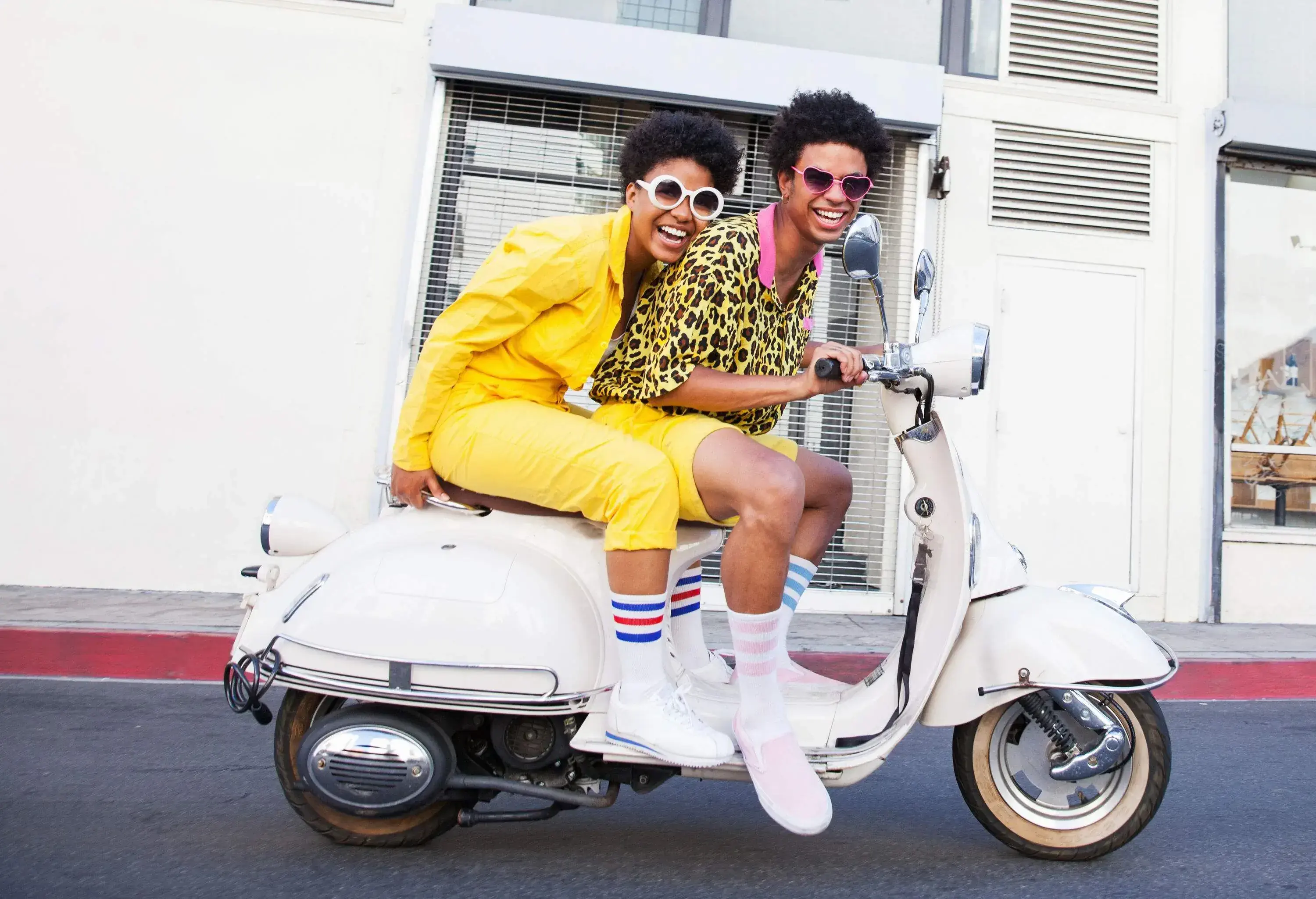 Two friends in vibrant yellow clothes, long white socks, and fashionable sunglasses ride a scooter on an asphalt street.