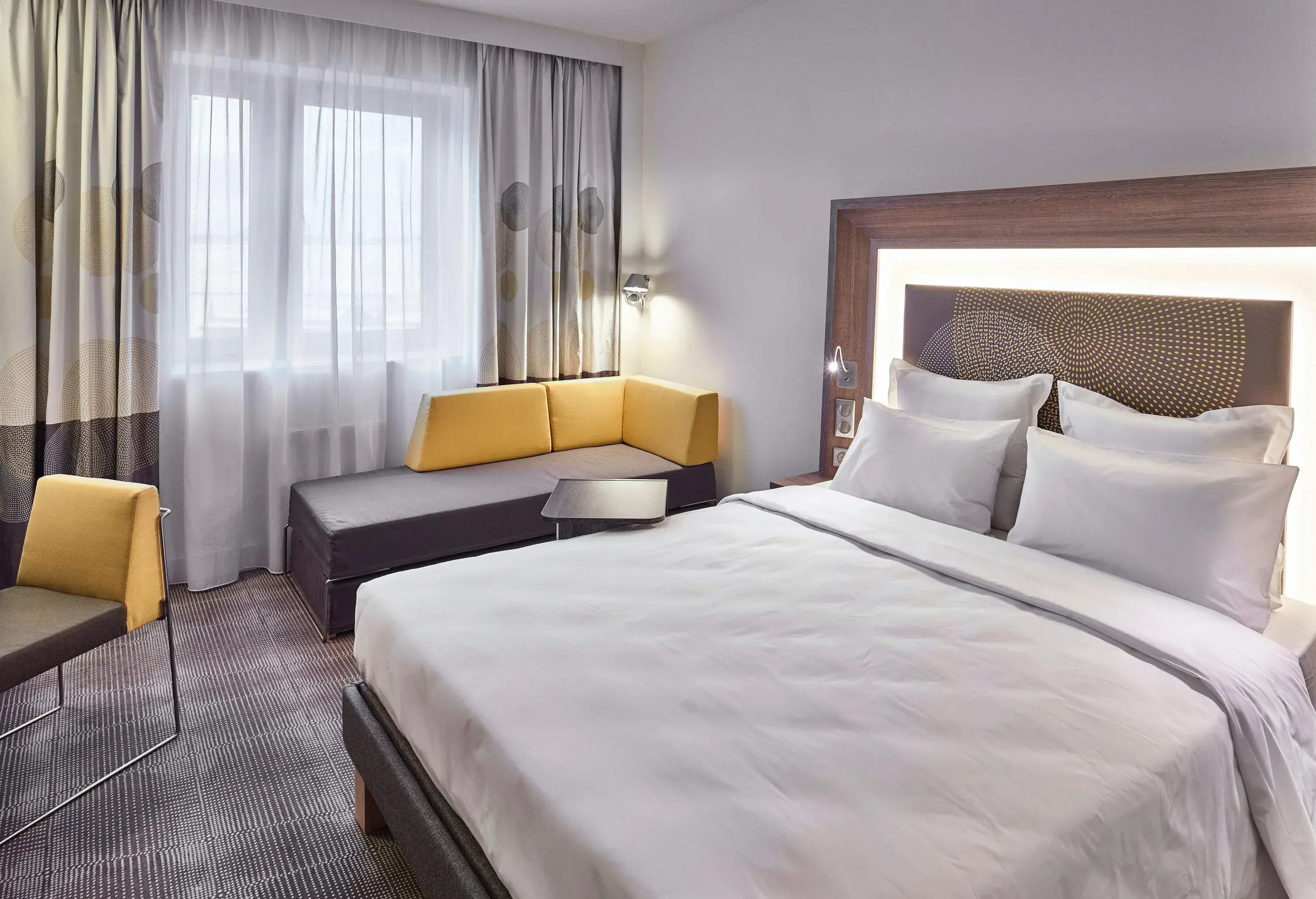 Interior of a hotel room with a big bed, a small couch, and a glass window behind the thin curtains.
