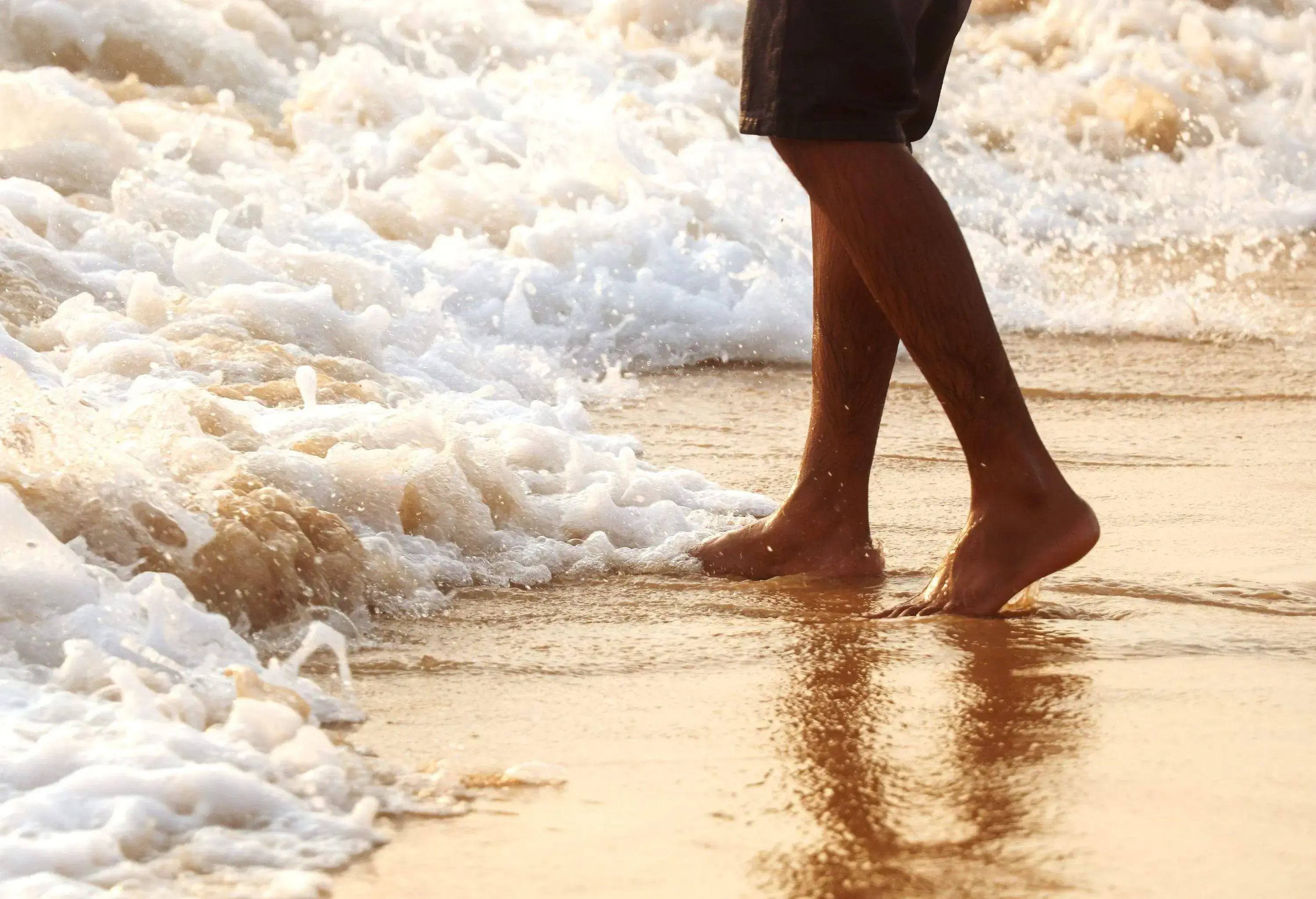 The white waves crashing into the sand with a man's feet standing.