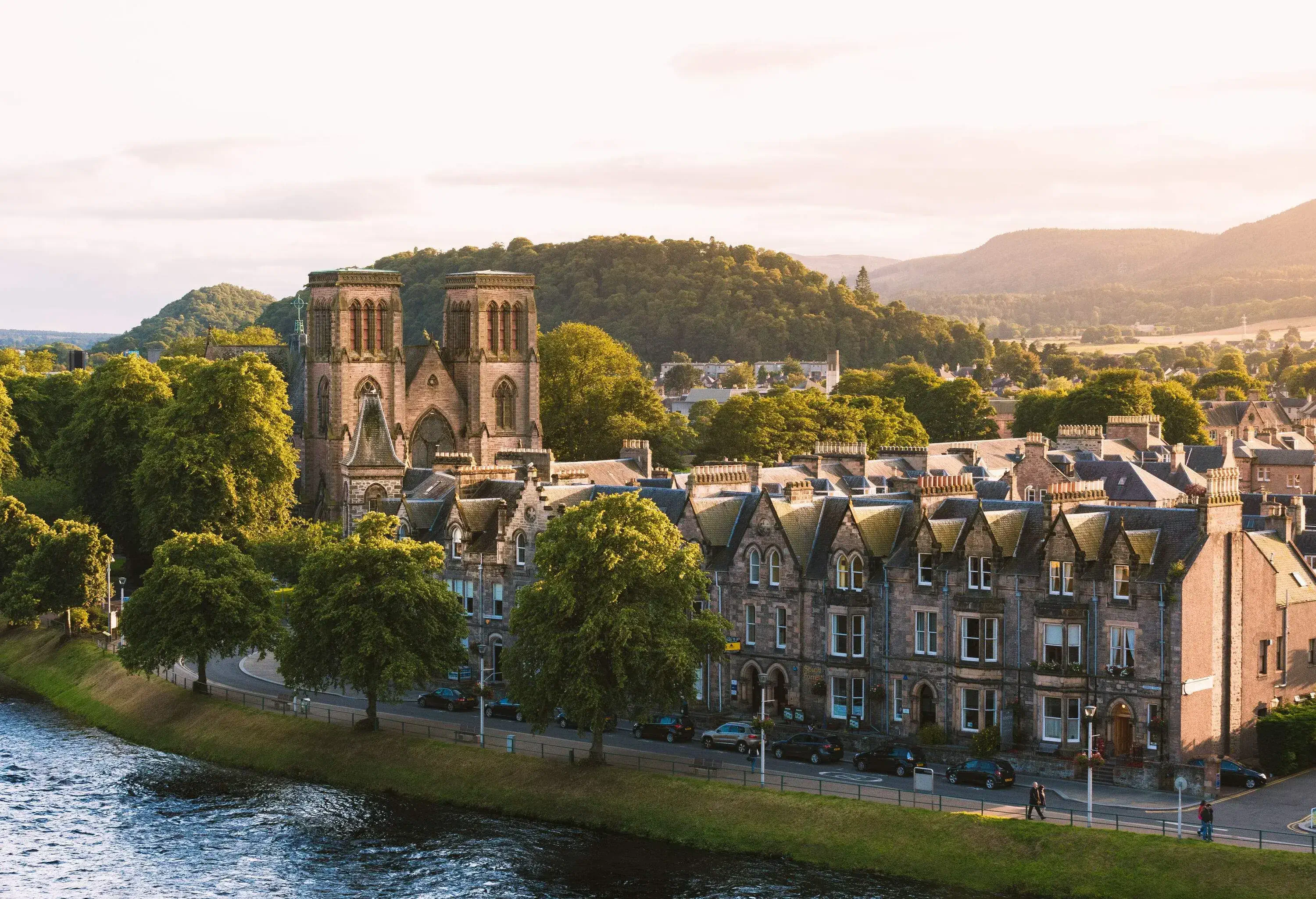 A brick church with twin towers overlooks the terraced buildings along a calm river.