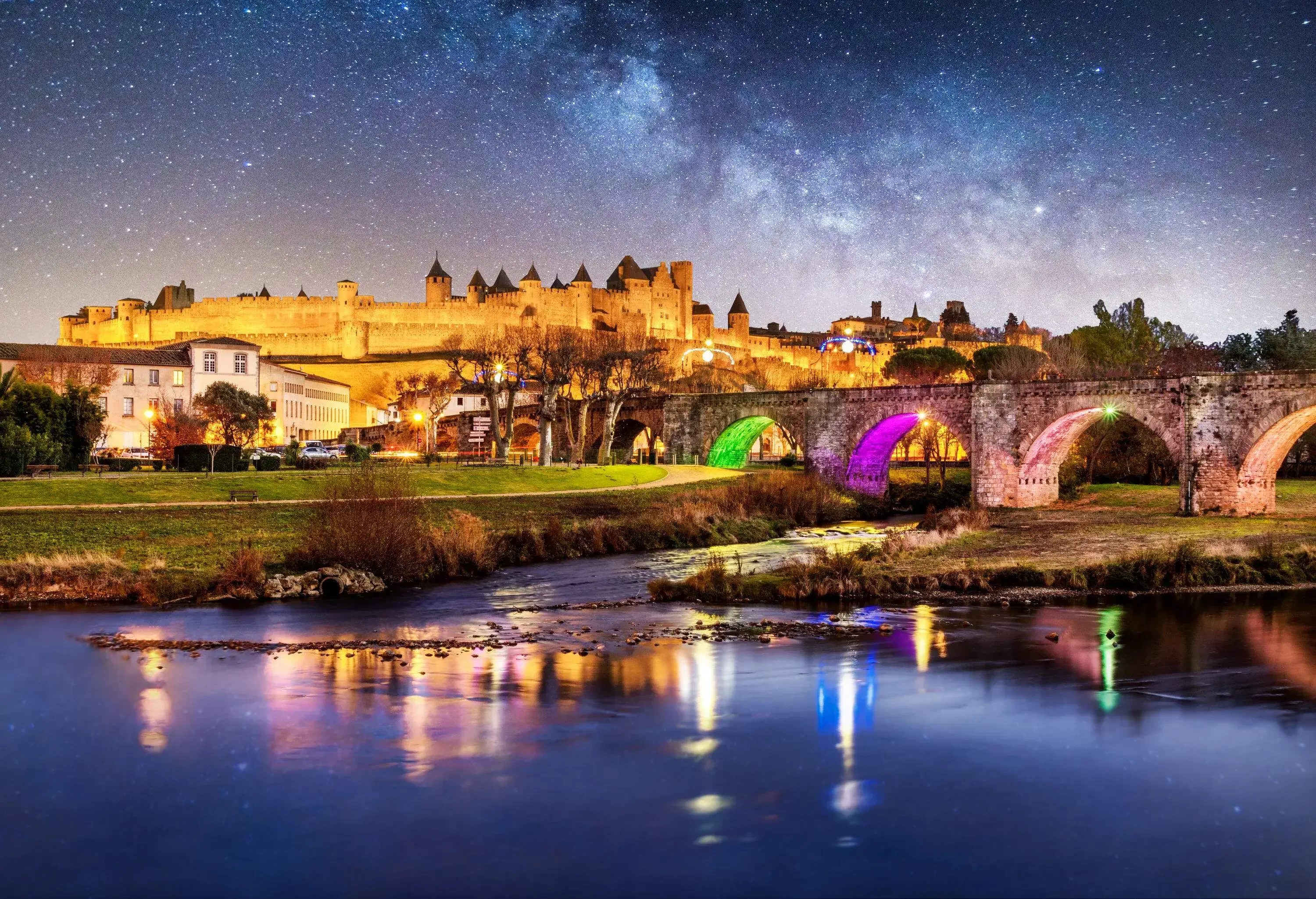 An old arch bridge across the river outside the thick wall of an illuminated castle at night.