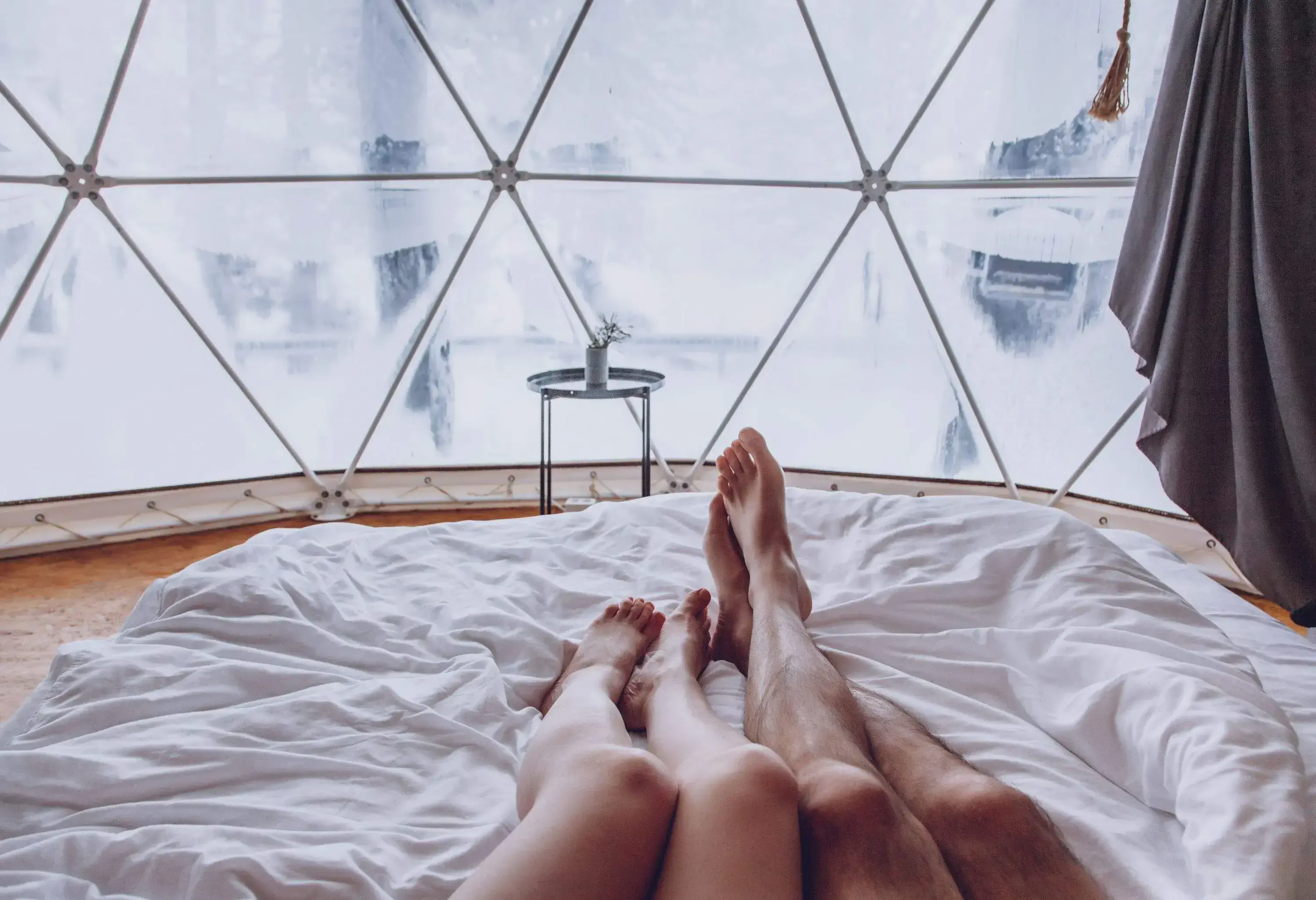 Legs of a man and woman couple in bed against the background of a snow-covered forest in a dome camping. Glamping vacation lifestyle concept.