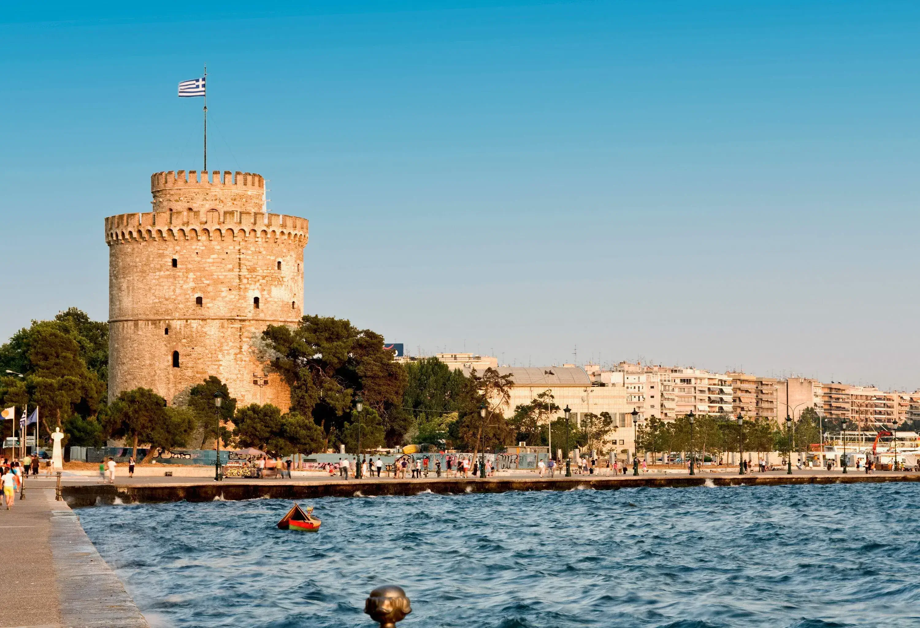 A white cylindrical tower with a Greece flag on a pole along a busy promenade.