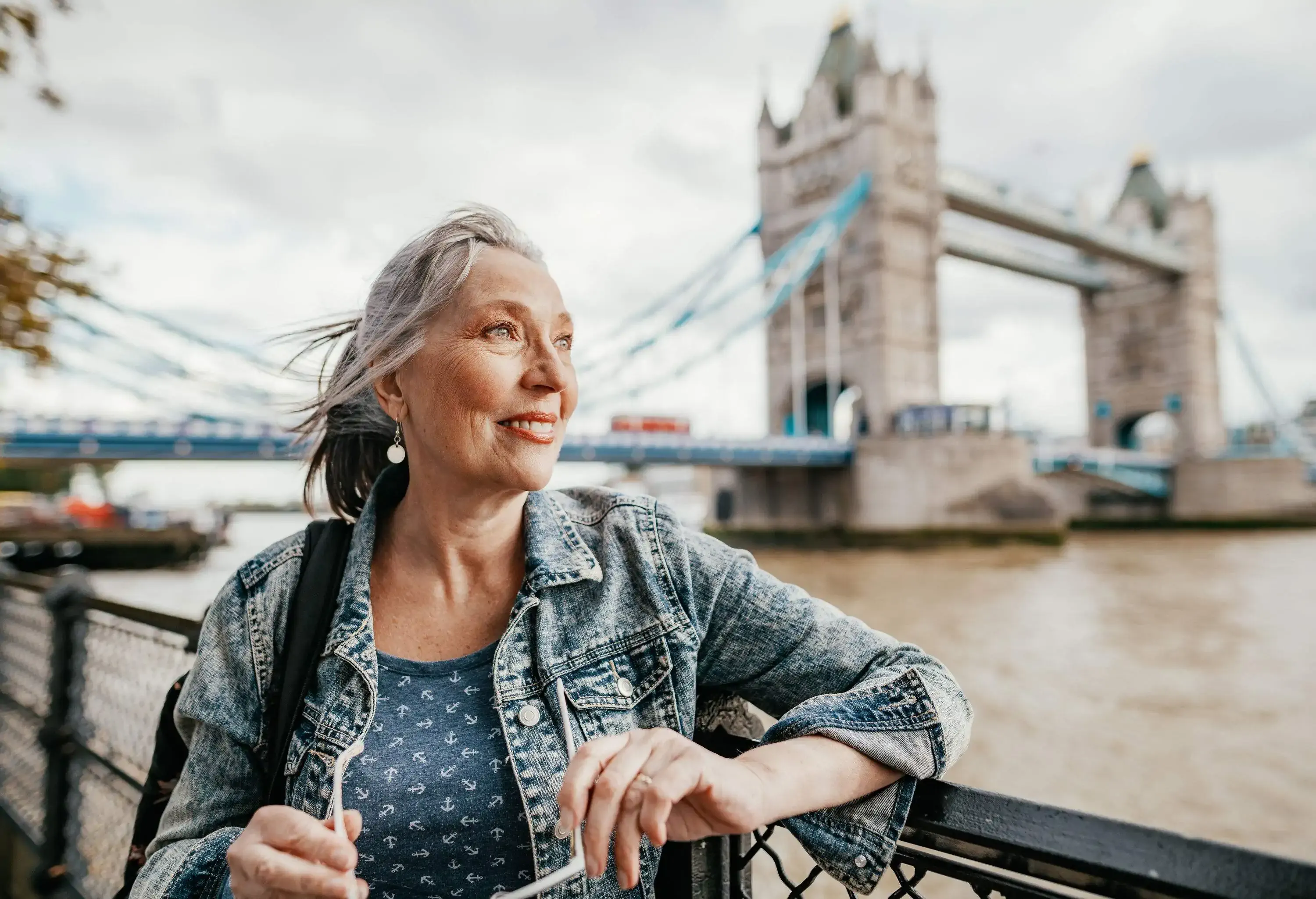 A senior woman leans on a fence with a background of the famous Tower Bridge across the river.