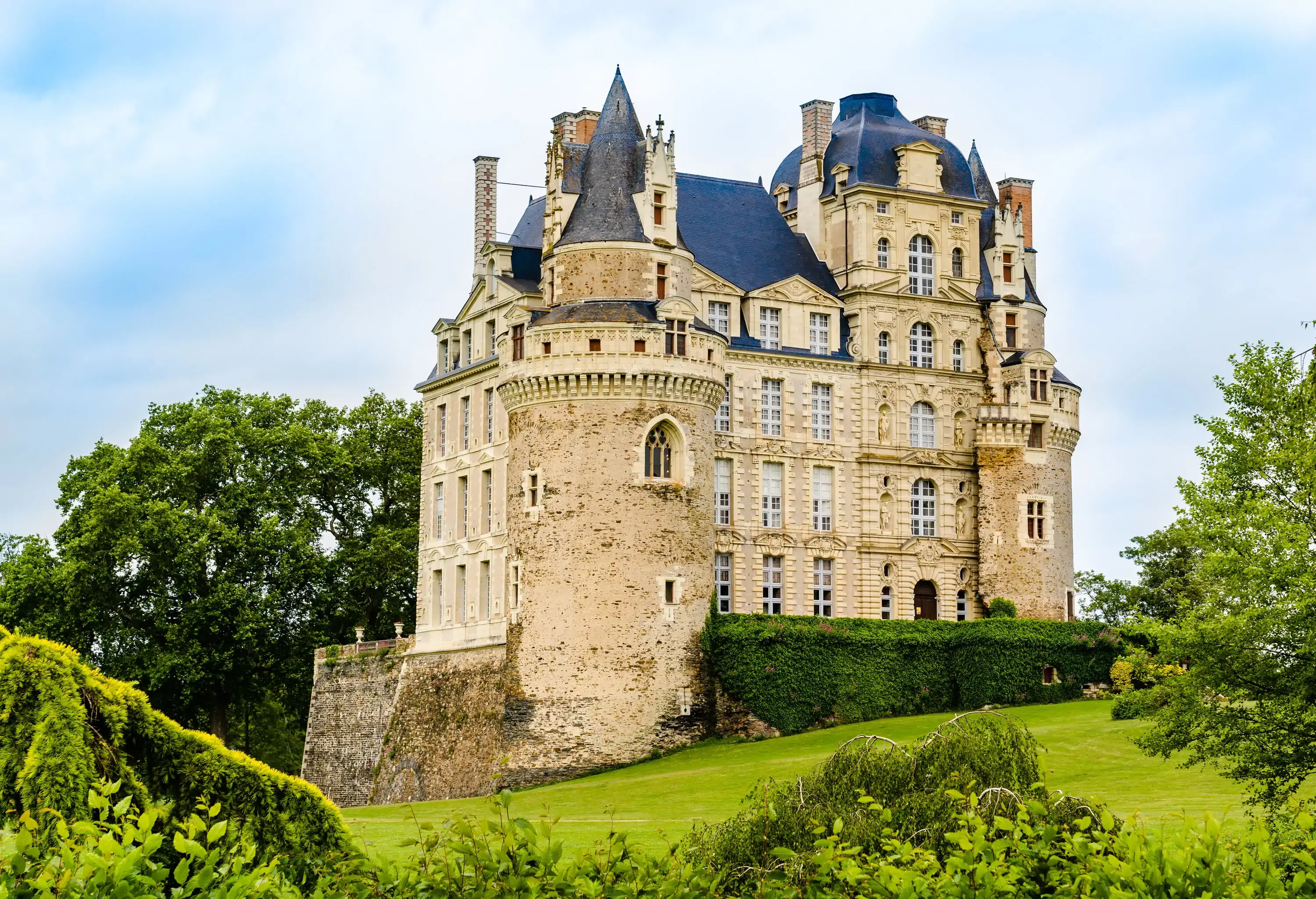 A Renaissance castle with a raised basement, round towers at each corner, and a pointed roof, all set in a lush, verdant landscape.