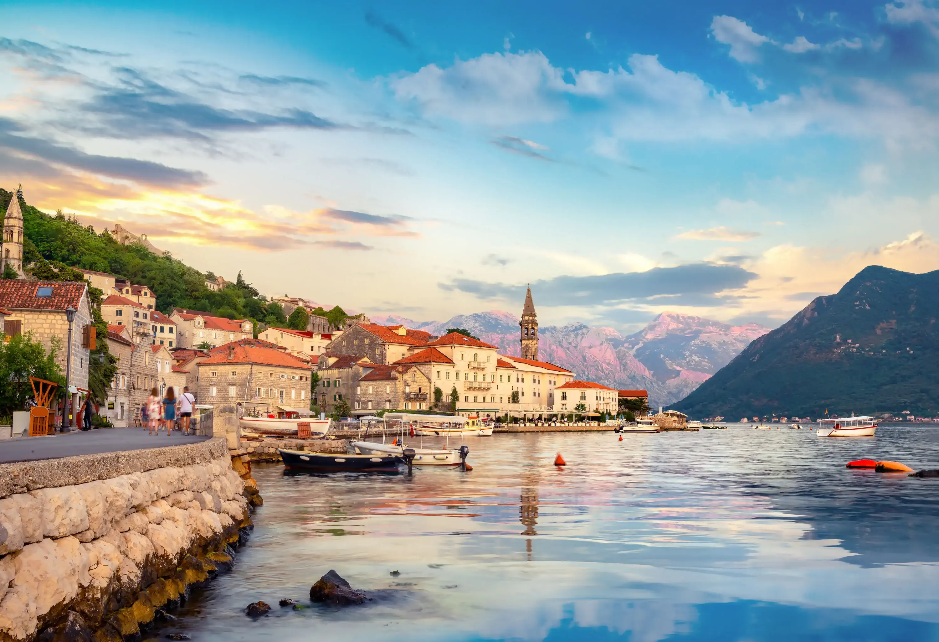 People stroll on a promenade along a bay with views of a coastal city with buildings perched on the hillsides.
