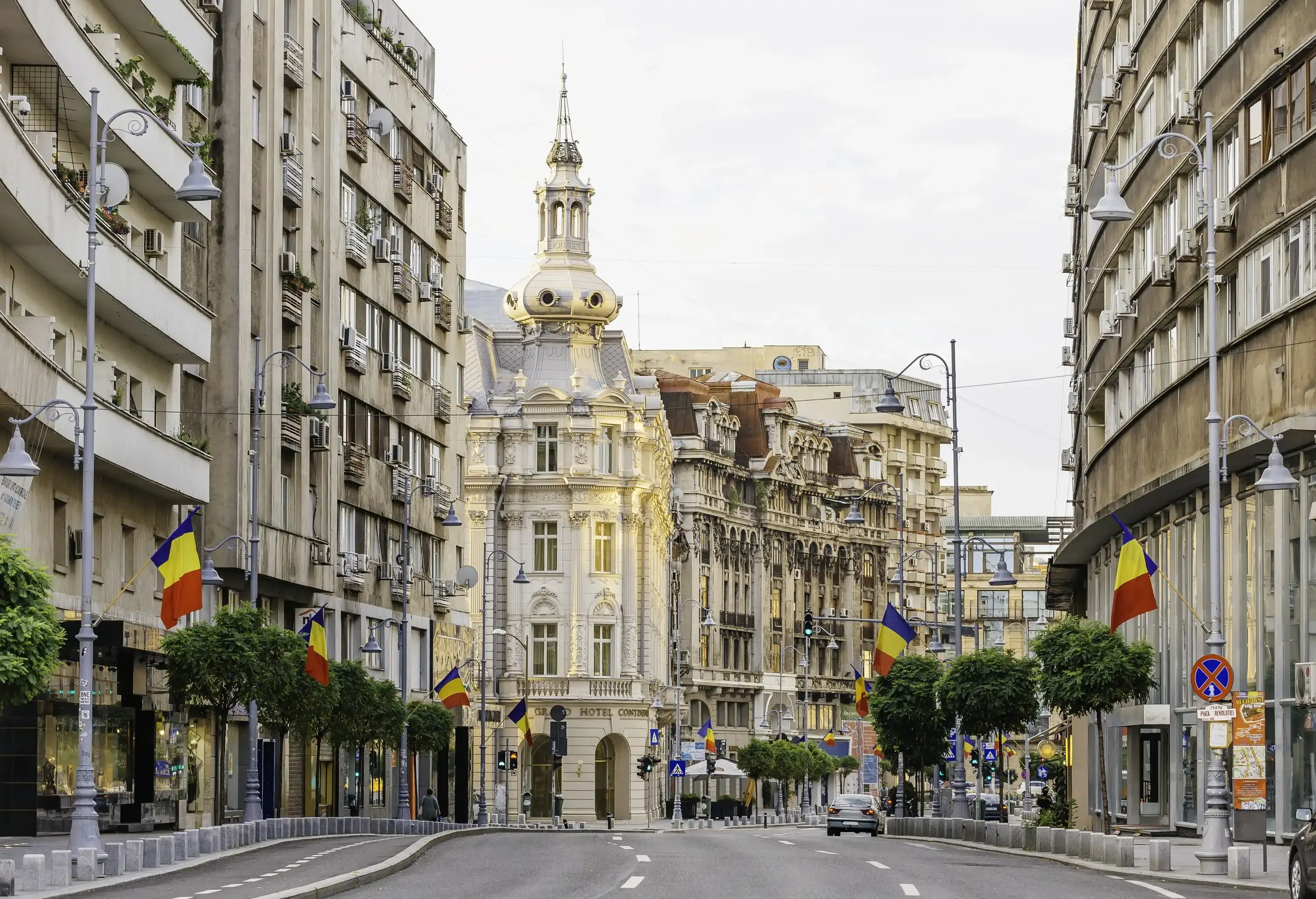 A tree-lined highway with cars bordered by old adjacent city buildings with waving Romanian flags.