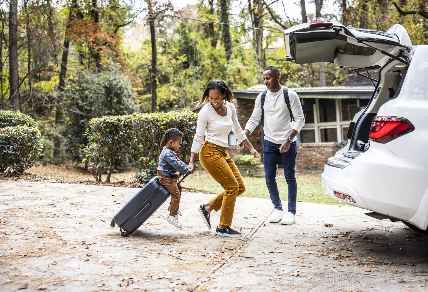 Mother pulling young boy on suitcase and loading car for family vacation